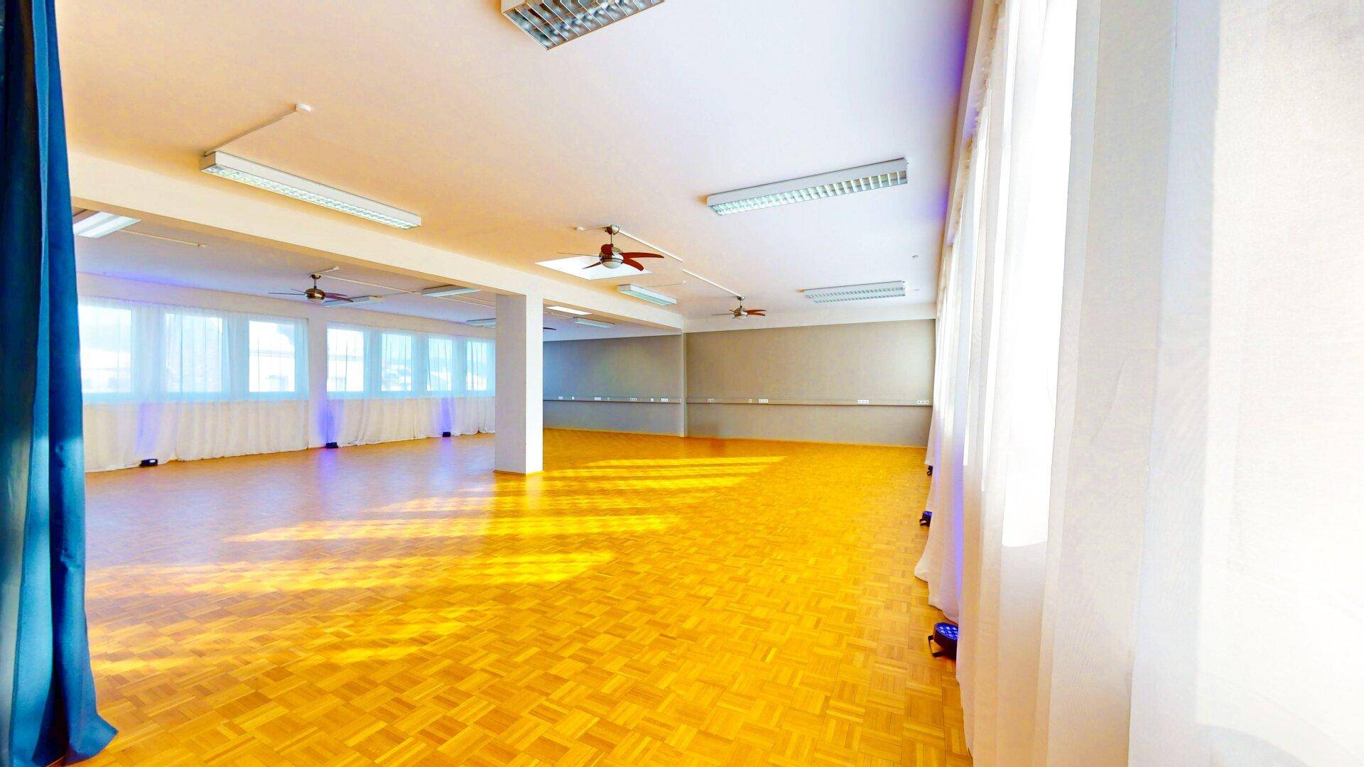 Geräumiger, heller Mehrzweckraum mit vielen Fenstern, Holzparkett und Deckenventilatoren, bereit für verschiedene Nutzungen.