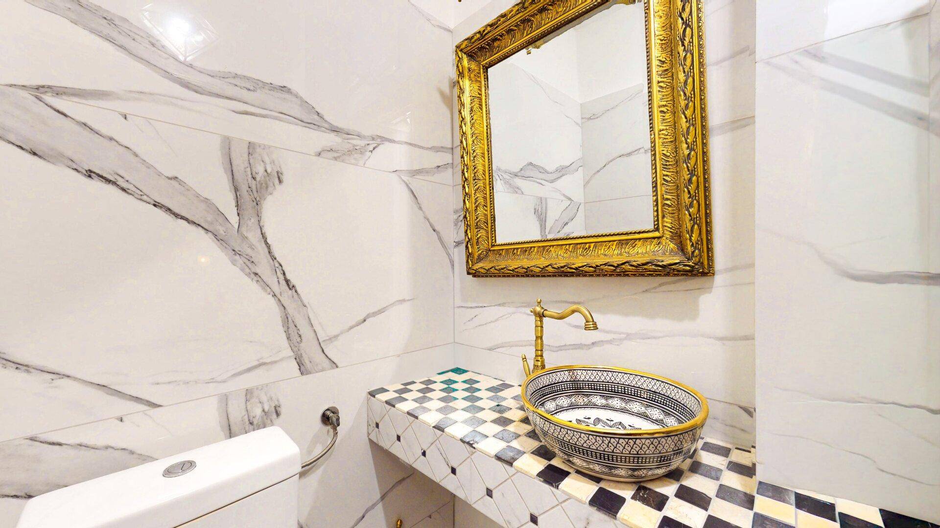 Elegantes Gäste-WC mit weißen Marmorwänden, dekorativem Waschbecken und goldenem Spiegel.