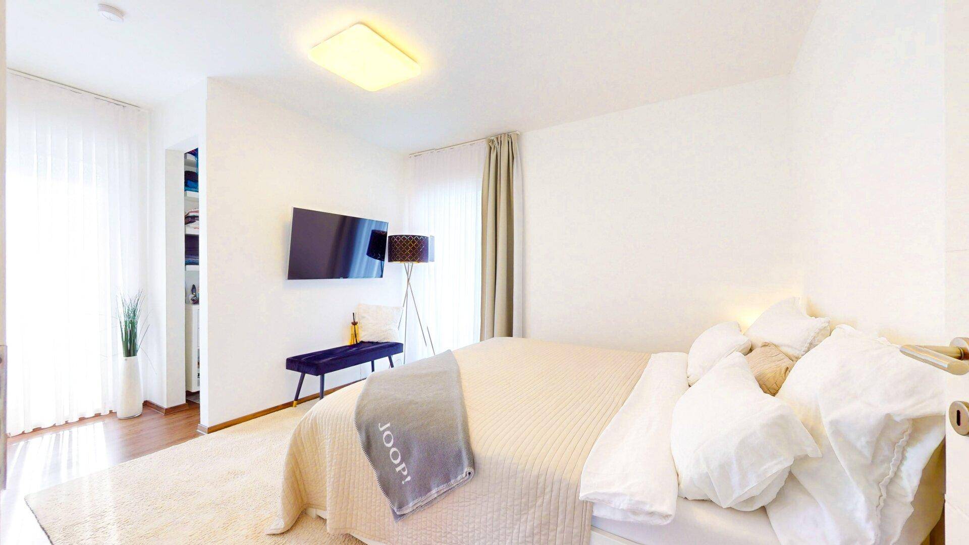 Helles Schlafzimmer mit Doppelbett, Wandfernseher und Zugang zu einem Balkon.