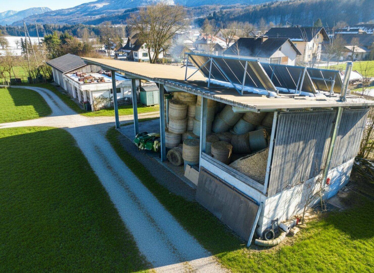 Nahaufnahme eines landwirtschaftlichen Gebäudes mit Solaranlagen auf dem Dach, umgeben von grünen Feldern.