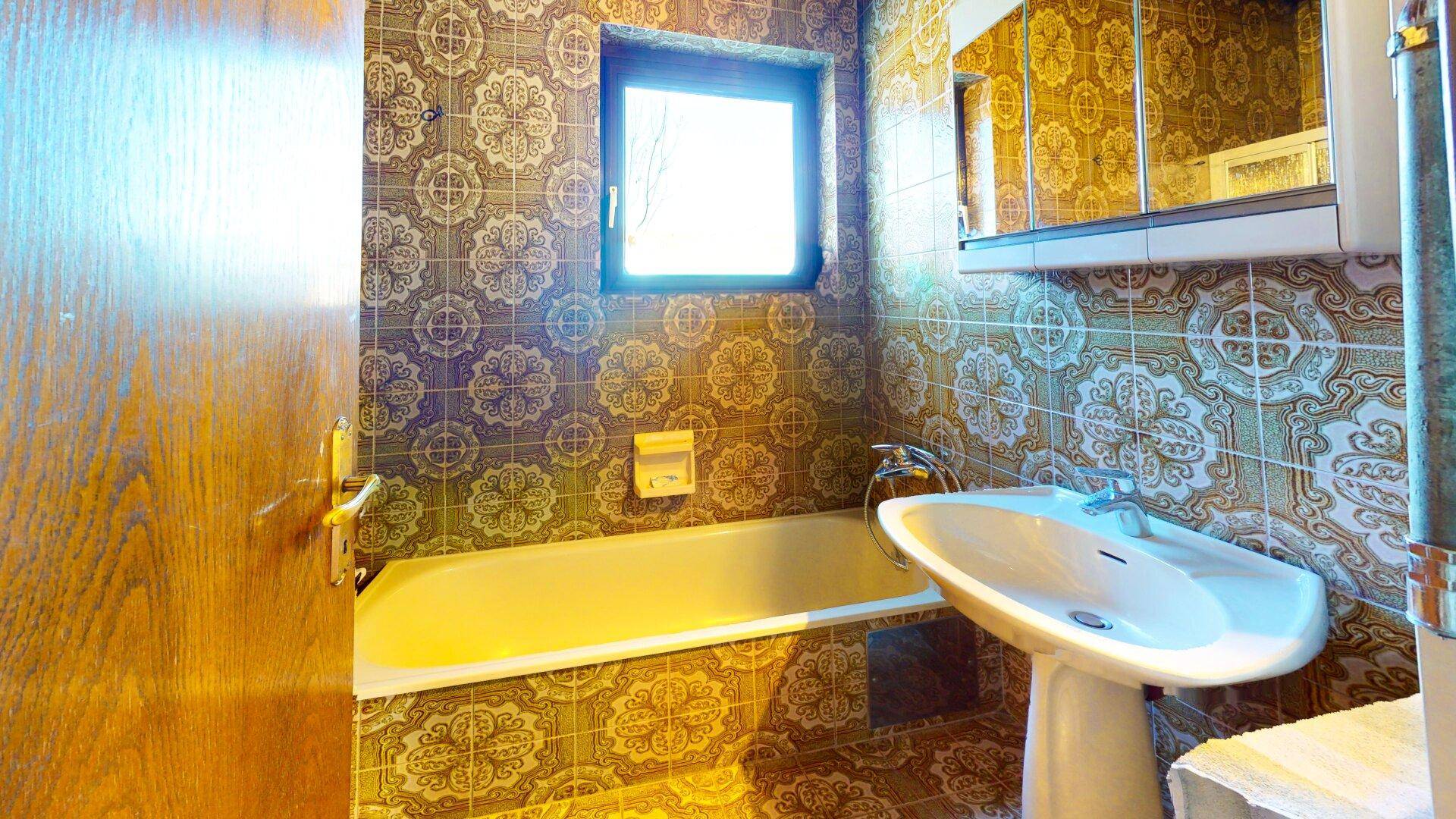 Badezimmer mit Badewanne, Waschbecken und Fenster, charakterisiert durch gemusterte Fliesen.