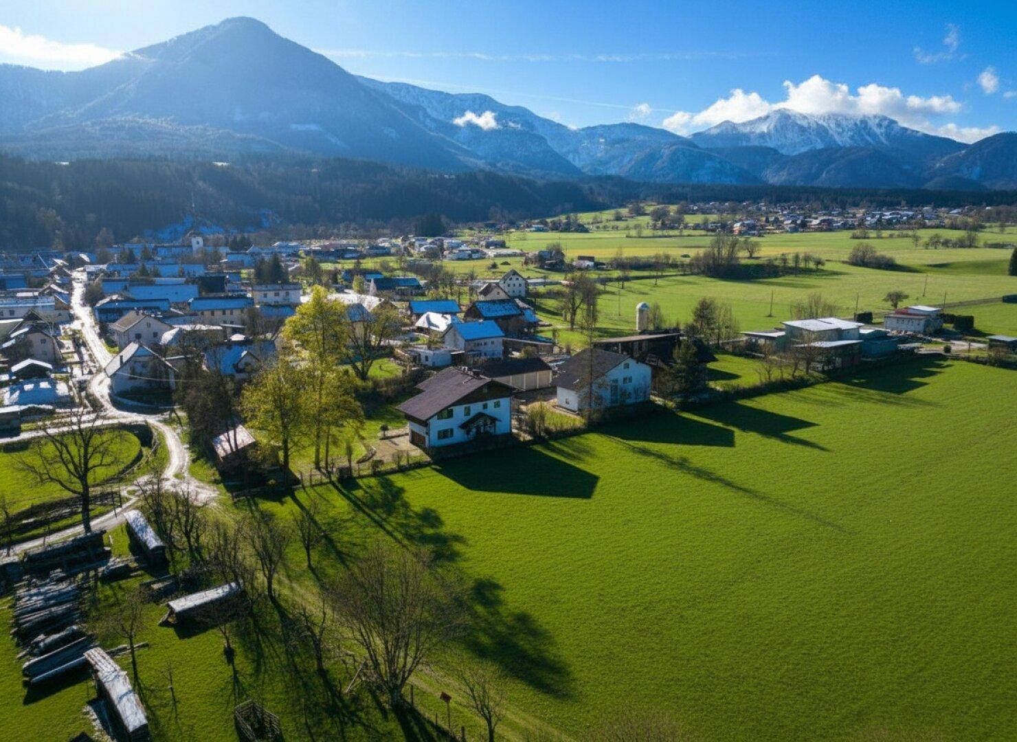 Luftaufnahme eines Dorfes eingebettet in grüne Felder mit schneebedeckten Bergen im Hintergrund.