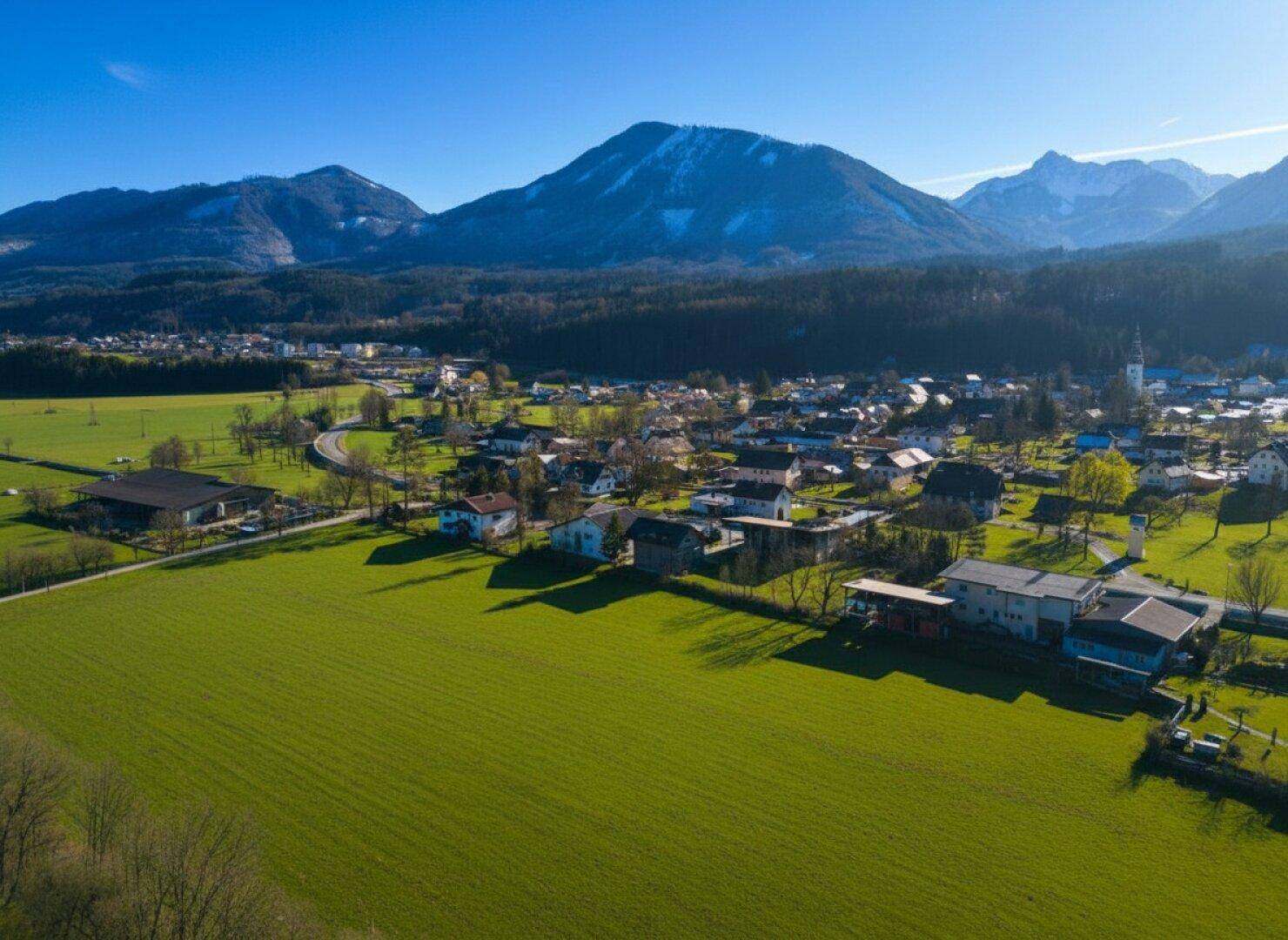 Weitläufige Luftaufnahme einer ländlichen Siedlung mit grünen Wiesen und imposanten Bergen.