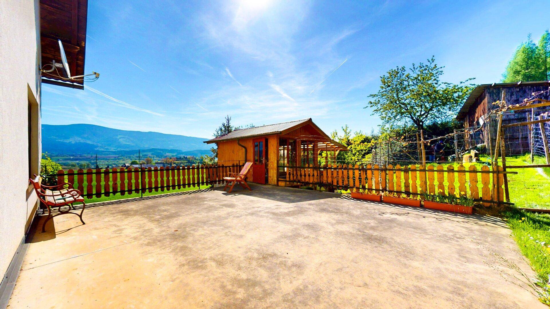 Sonniger Terrassenbereich mit Gartenhaus, Holzzaun und weitem Blick auf die Berge.