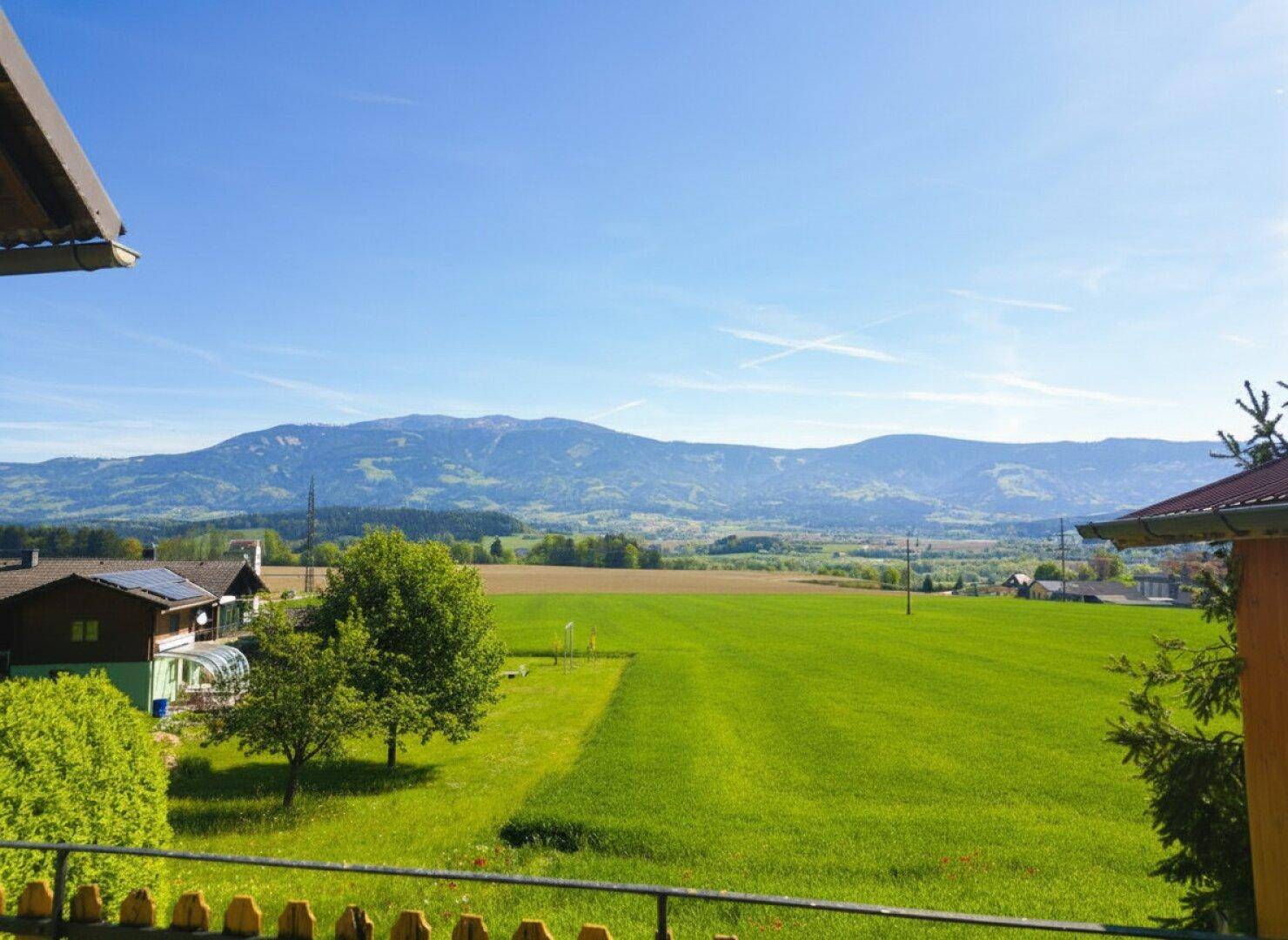 Weitläufiger Blick über grüne Felder und sanfte Hügel mit Bergen im Hintergrund unter blauem Himmel.