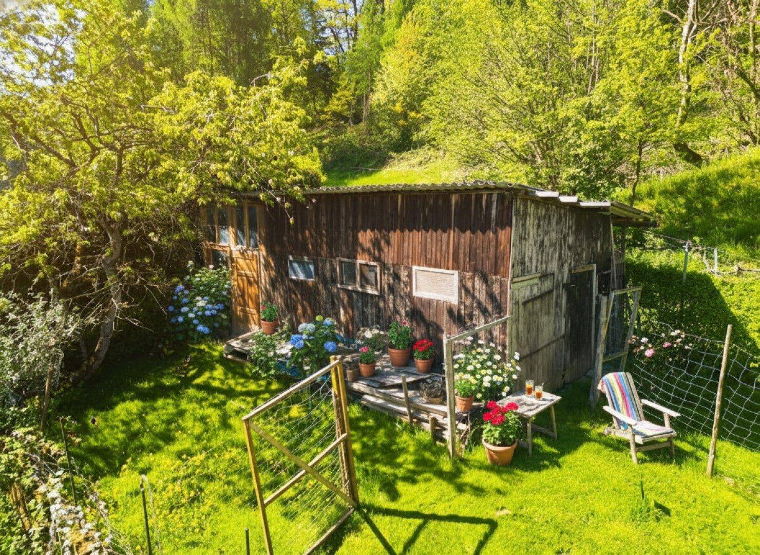 Rustikales Gartenhaus aus Holz, umgeben von Blumen und grüner Wiese in sonniger Lage.
