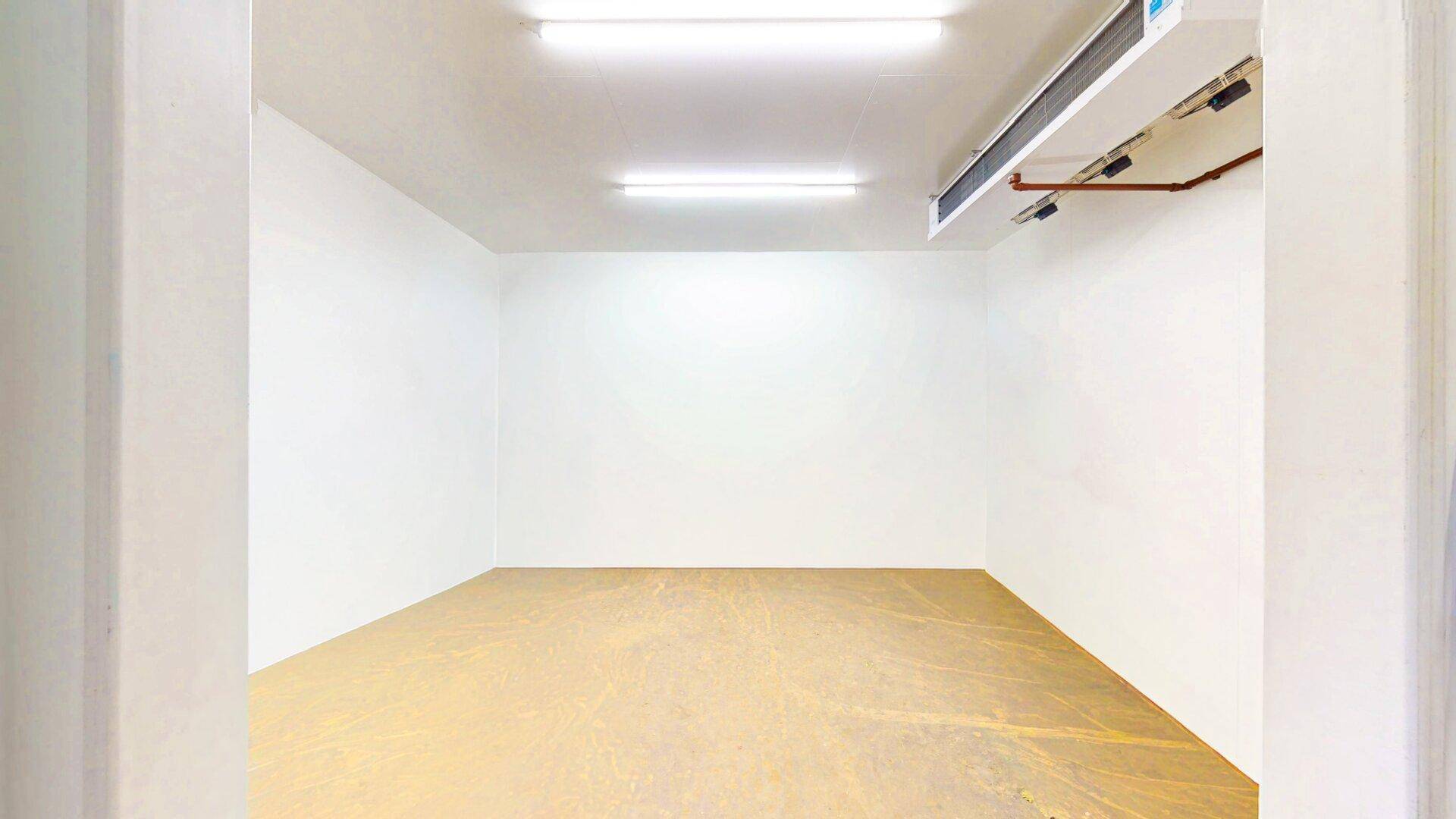 Leerer Kühlraum oder Vorratsraum mit weißen Wänden, hellem Boden und sichtbaren Lüftungsrohren an der Decke.