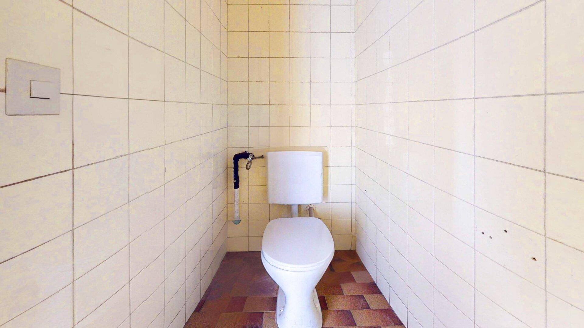 Einfache Toilette mit weißen Fliesen an den Wänden und braunen Fliesen auf dem Boden.