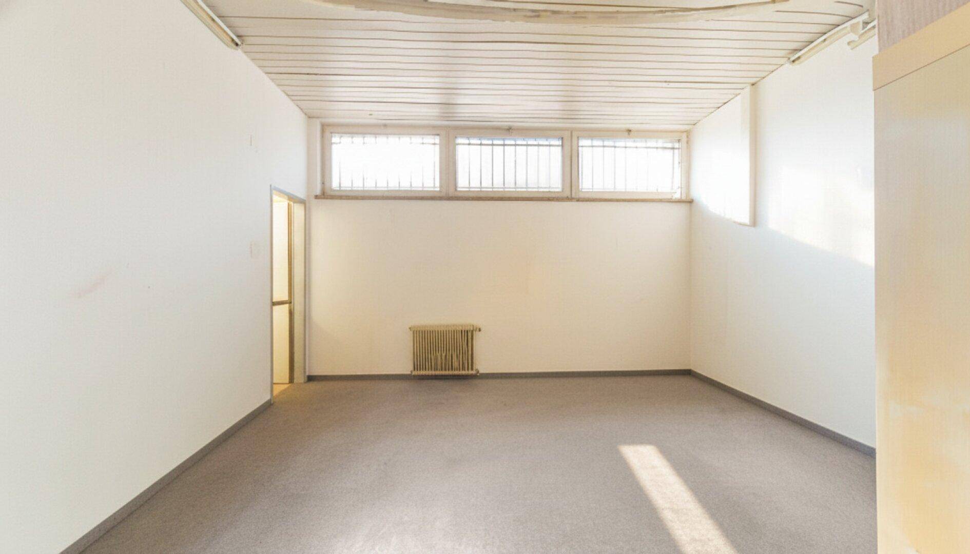 Leerer Büroraum mit grauem Teppichboden, weißen Wänden, kleinen Fenstern mit Gittern und einem Heizkörper.