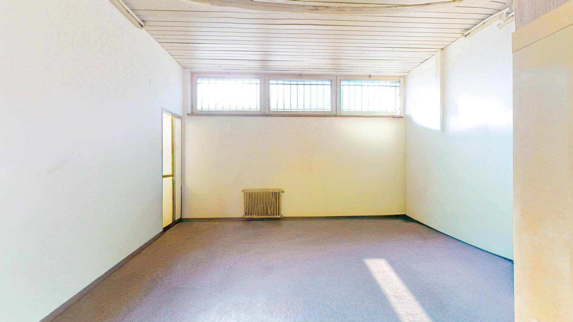 Leerer Raum mit hellen Wänden, grauem Boden, Heizkörper und hohen Fenstern mit Gitter.