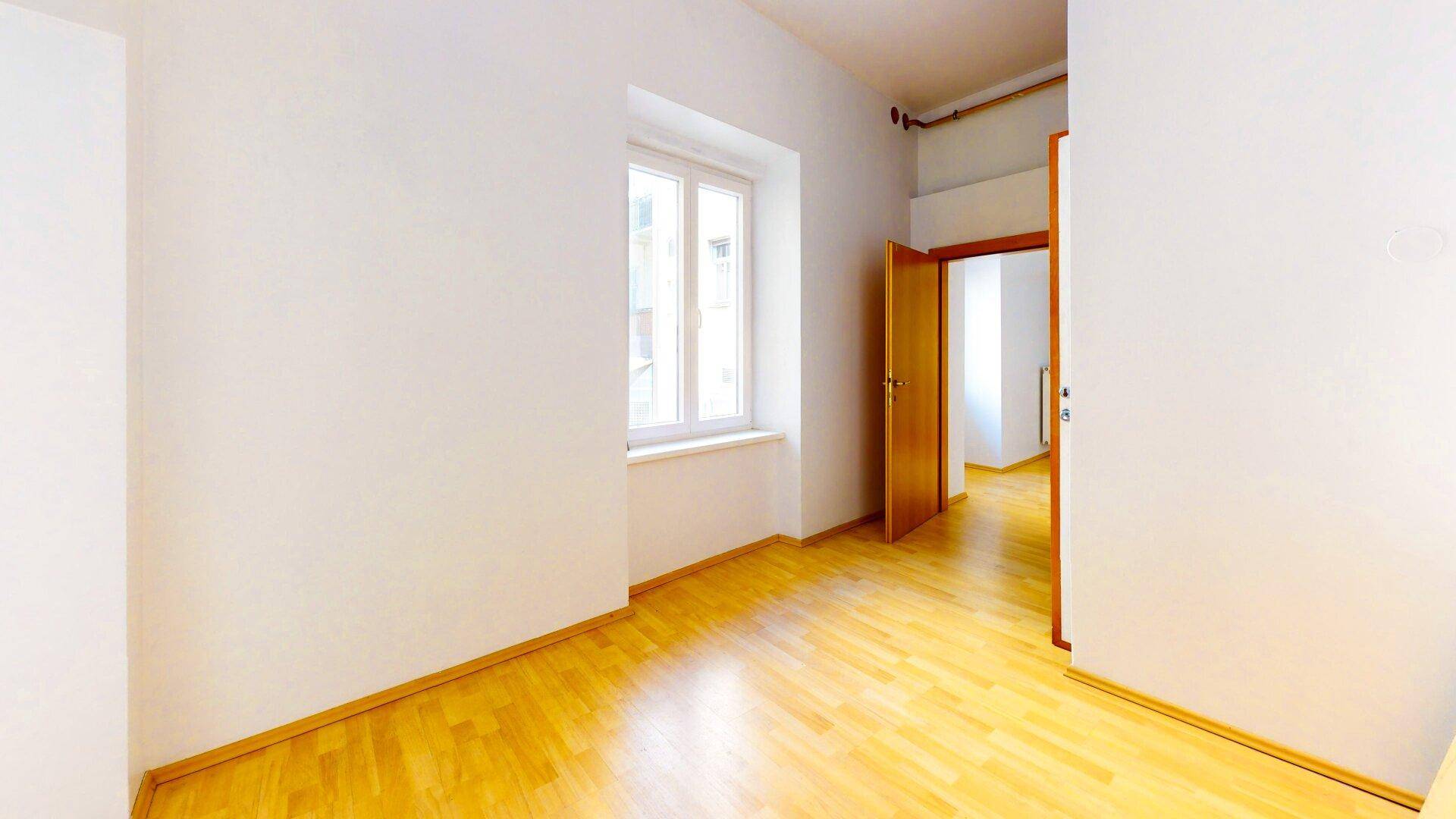 Leerer Raum mit Holzboden, weißen Wänden und einem Fenster, das Tageslicht hereinlässt.