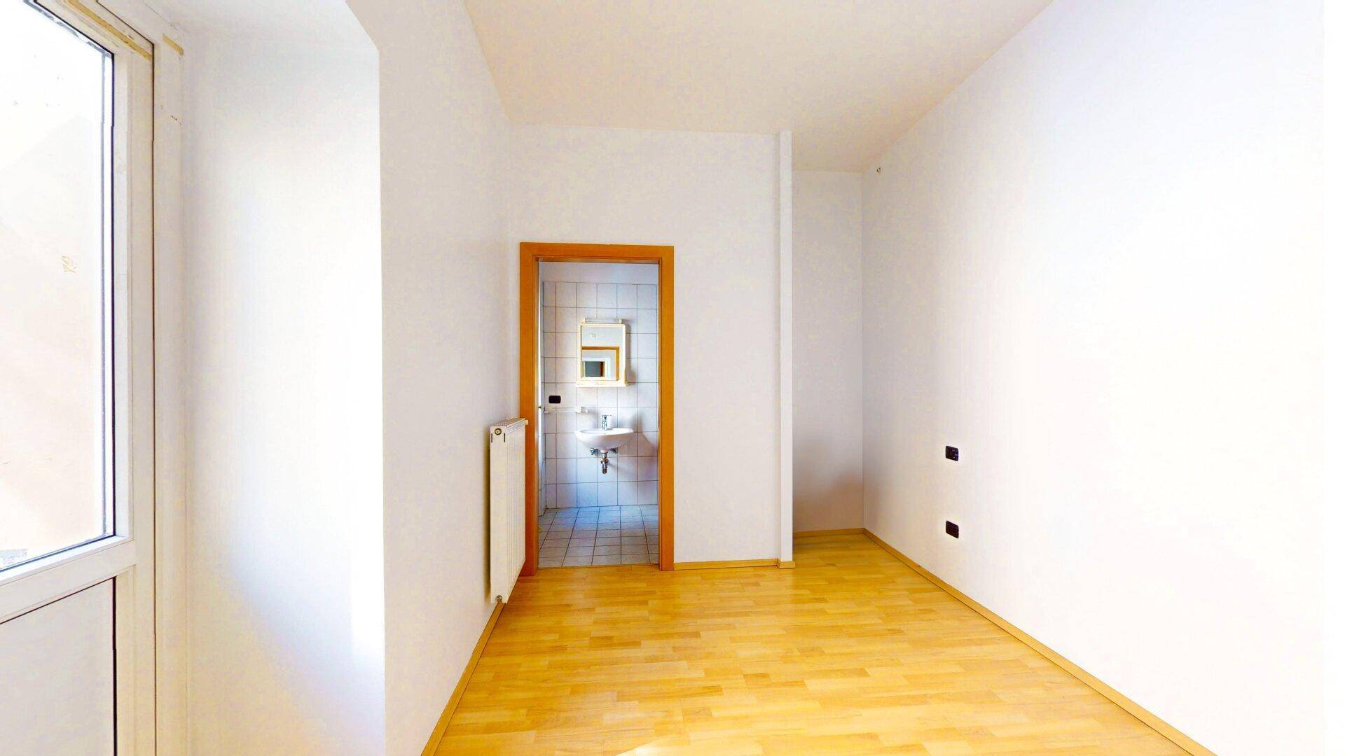 Leerer Raum mit Holzboden, weißen Wänden und Blick auf ein Badezimmer durch die offene Tür.