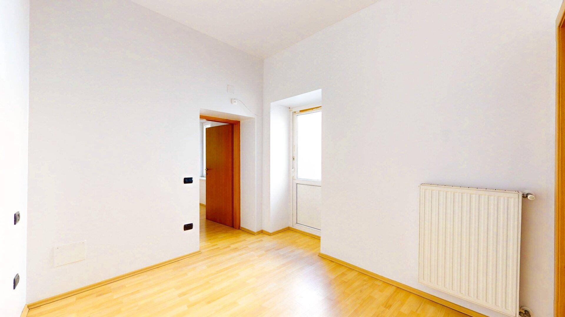 Heller Raum mit Holzboden, weißen Wänden, Heizkörper und einer Tür, die zu einem Balkon führt.