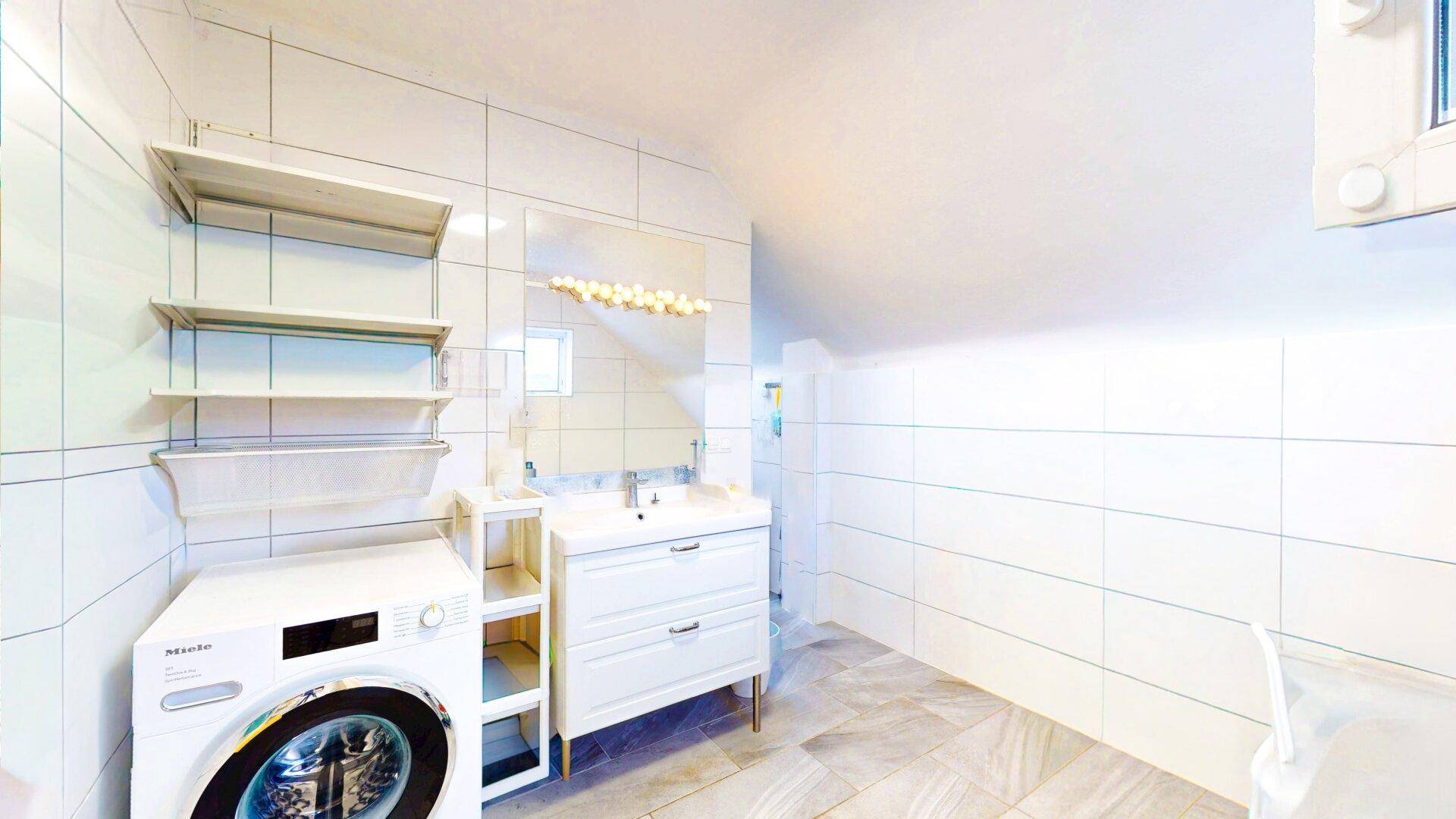 Helles Badezimmer mit Waschmaschine, Waschbecken und Spiegelschrank unter der Dachschräge.