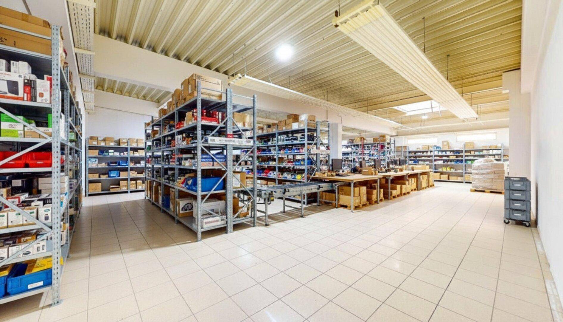 Weitläufiger Lagerraum mit Regalsystemen, Packstation und Förderband auf sauberem Fliesenboden.