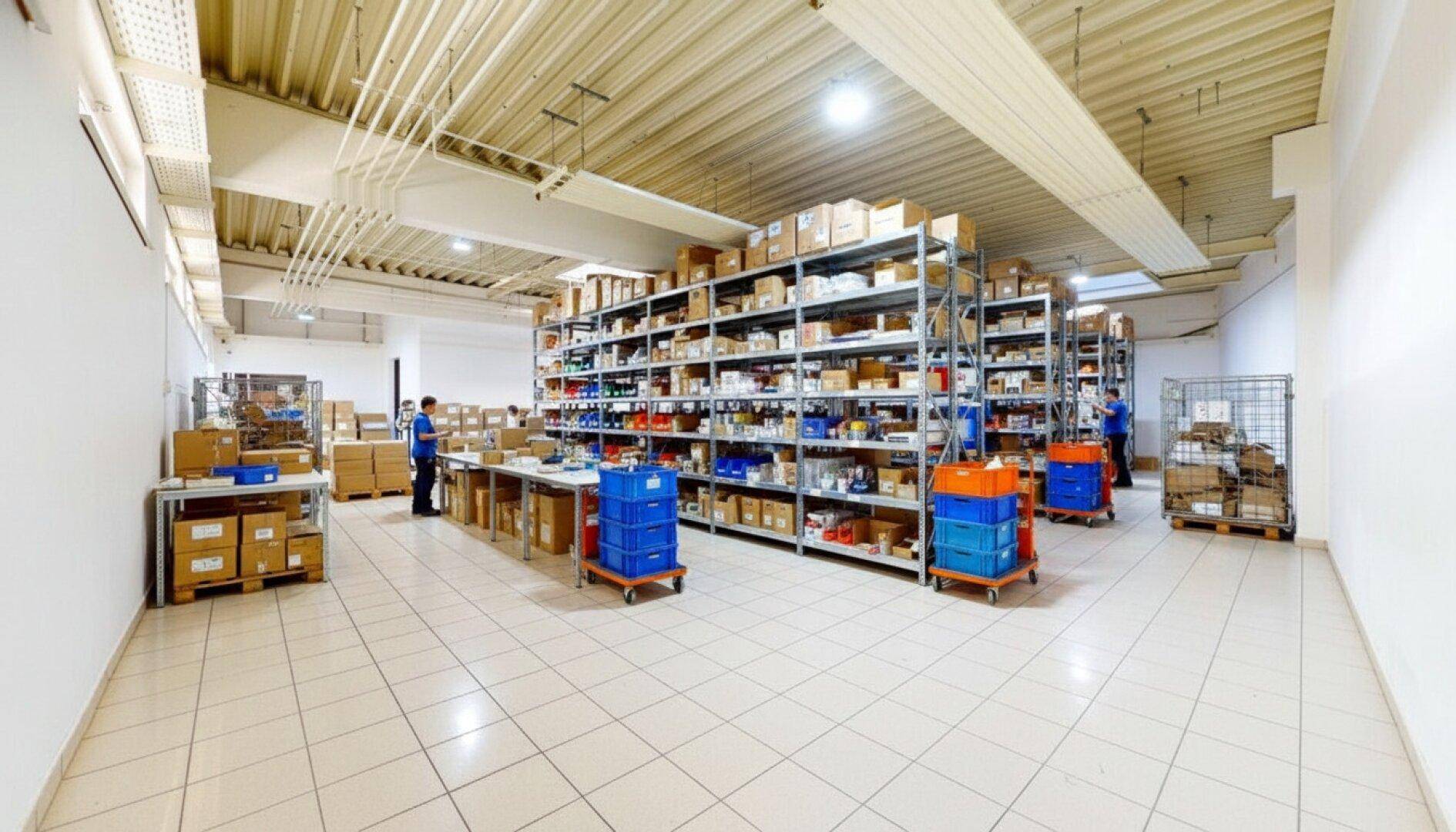 Lagerbereich mit Regalen, Arbeitstischen und mobilen Rollwagen, beleuchtet von Deckenleuchten.