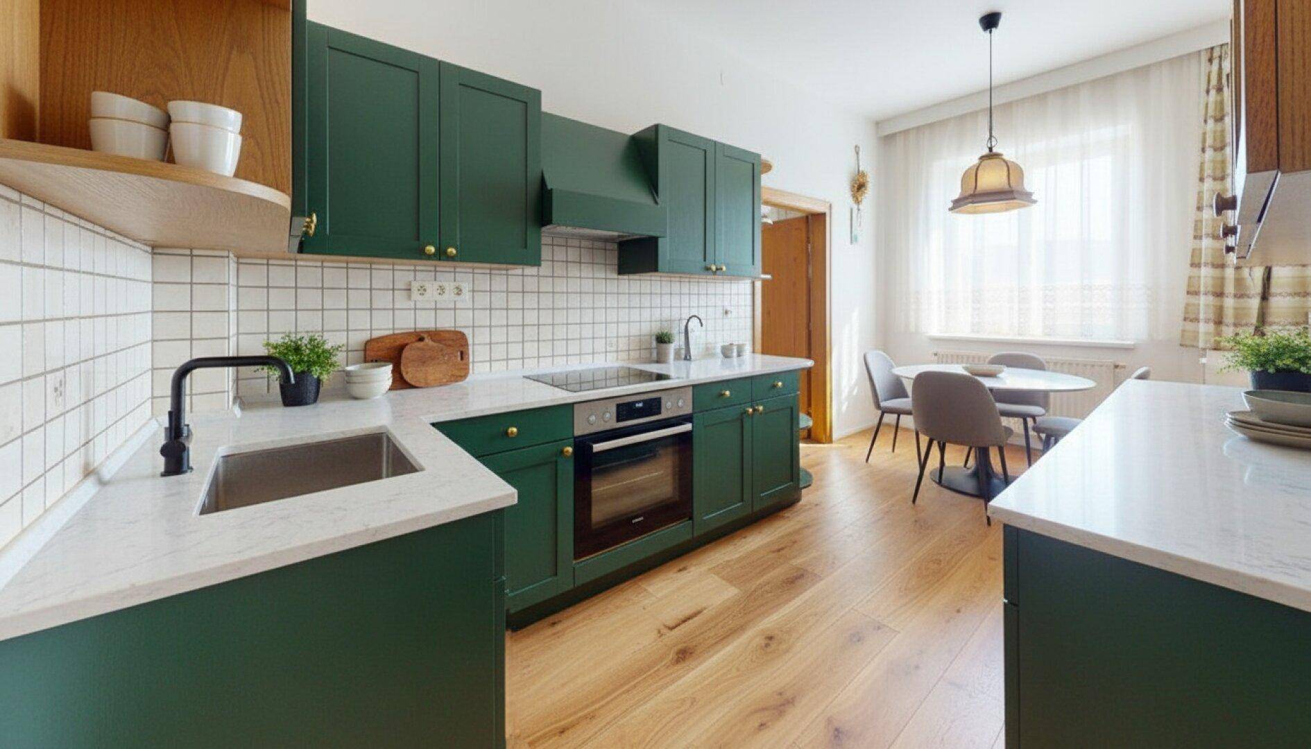 Moderne Küche in Dunkelgrün mit Holzboden, weißen Arbeitsplatten und Essbereich am Fenster.