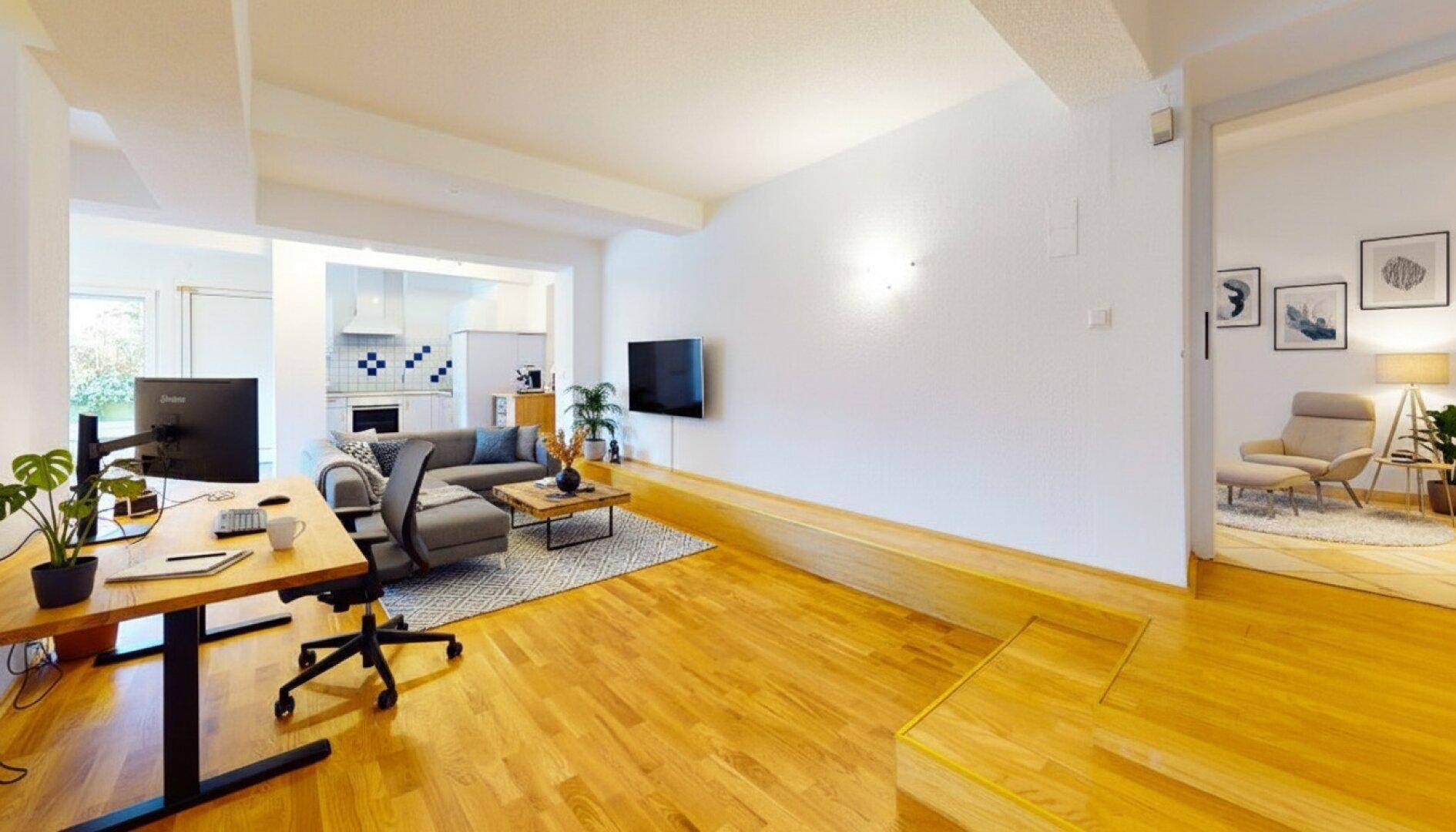 Großzügiger Wohnbereich mit Holzboden, integriertem Arbeitsplatz und Blick auf die offene Küche.