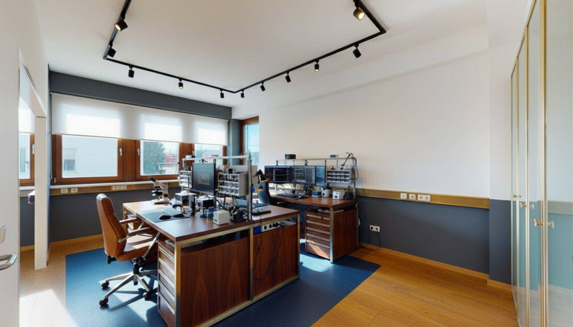 Büro mit mehreren Arbeitsplätzen, dunklen Holzschreibtischen und Schienenspots an der Decke.