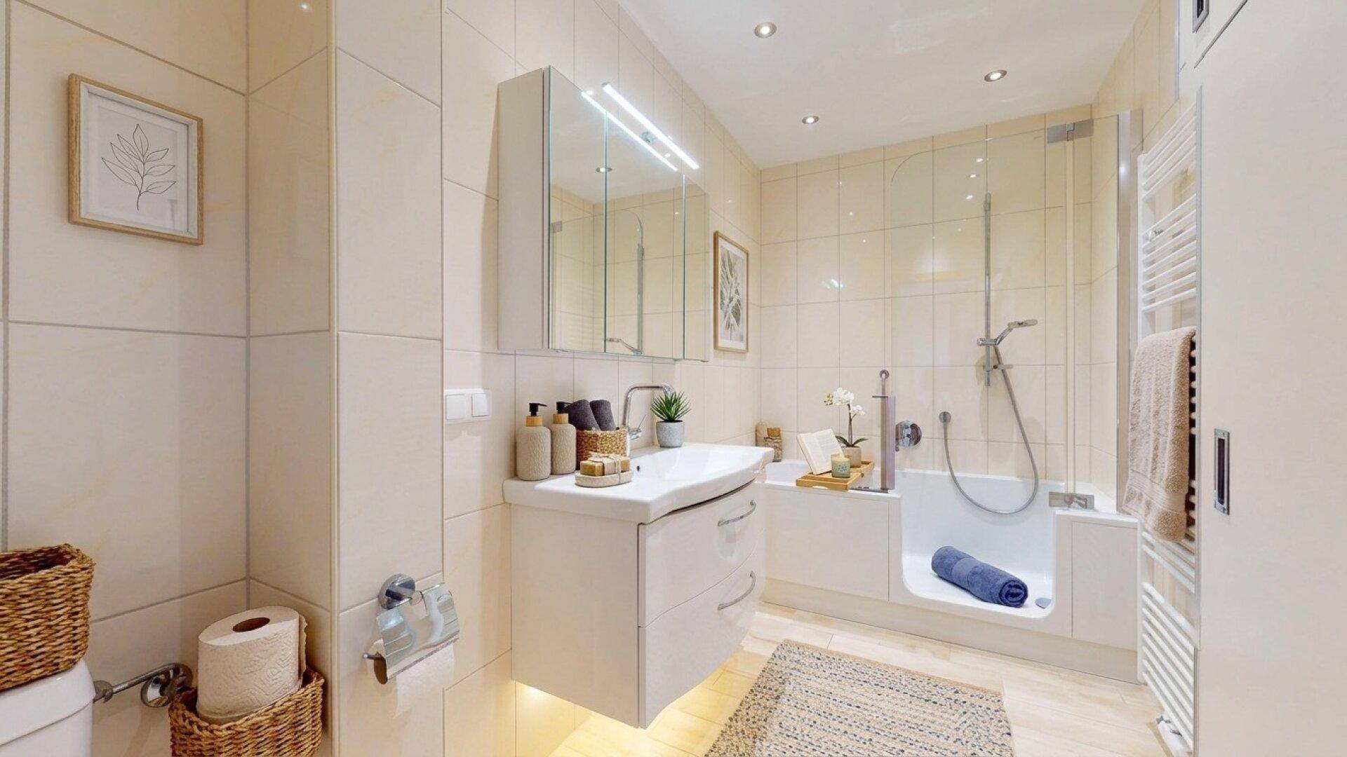 Freundliches Badezimmer mit beigefarbenen Fliesen, Waschtisch, Spiegel und Badewanne mit Duschfunktion.
