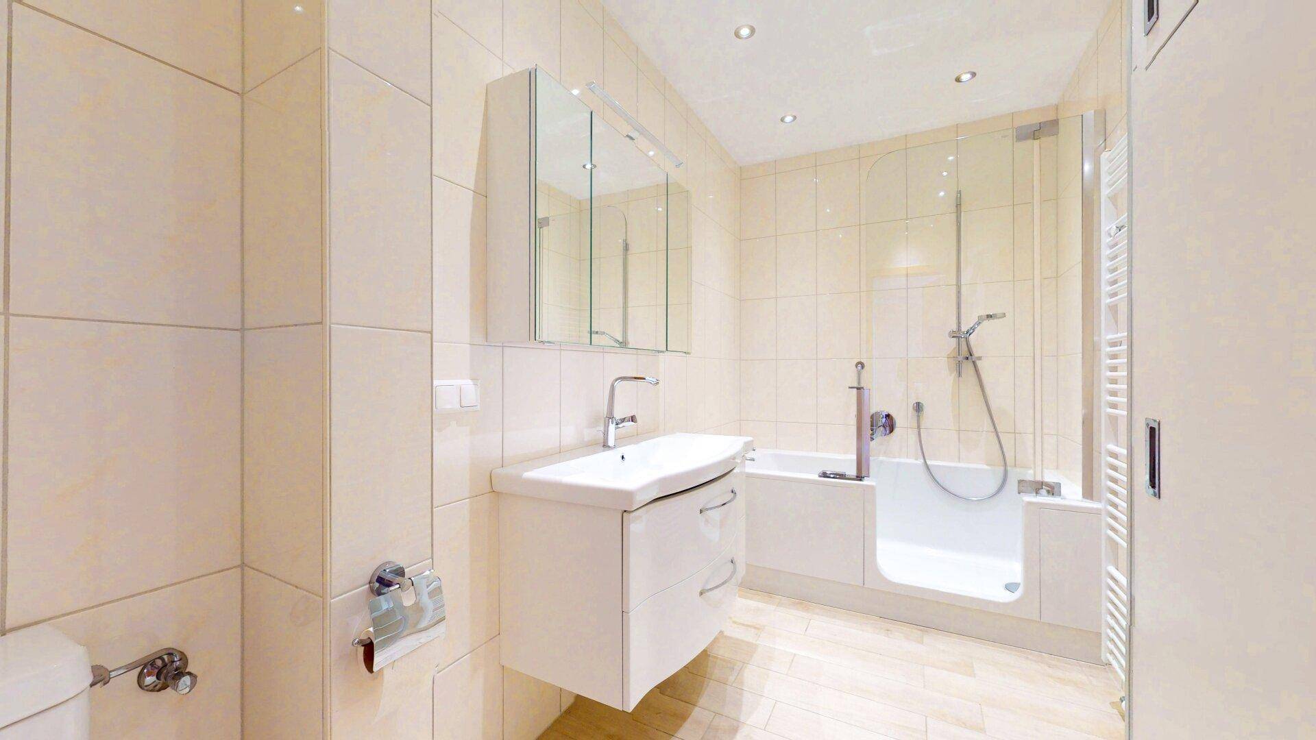 Helles Badezimmer mit beigefarbenen Fliesen, Waschtisch, Spiegelschrank und Badewanne mit Duschfunktion.