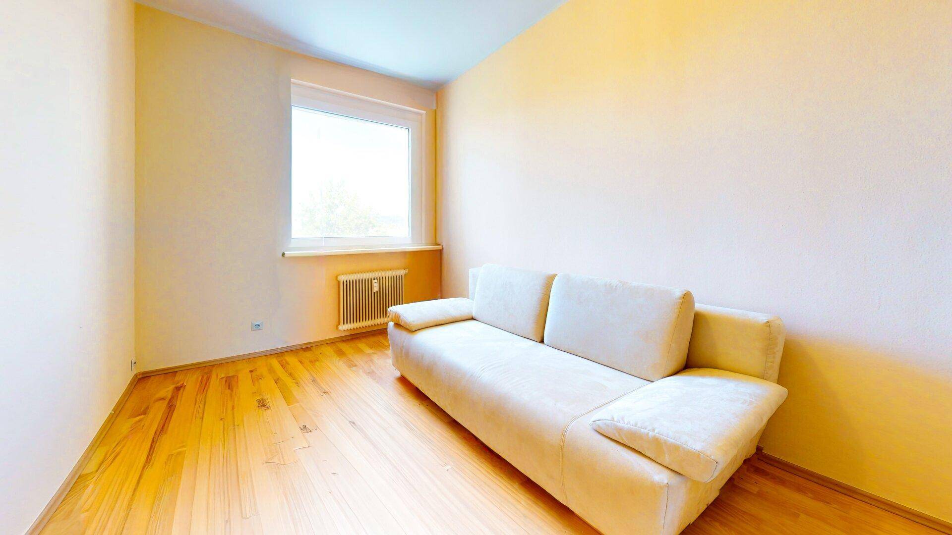 Helles Zimmer mit Holzboden, einem Sofa und Fenster, ideal als Schlaf- oder Gästezimmer nutzbar.