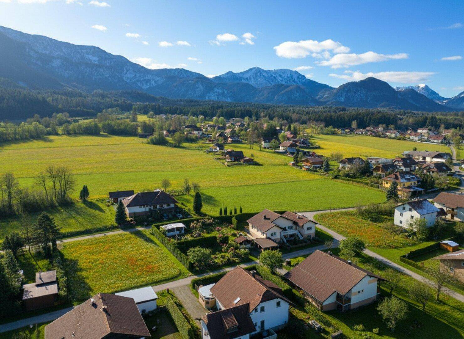 Weitläufige Landschaft mit Wohnsiedlung, grünen Feldern und majestätischen Bergen im Hintergrund.