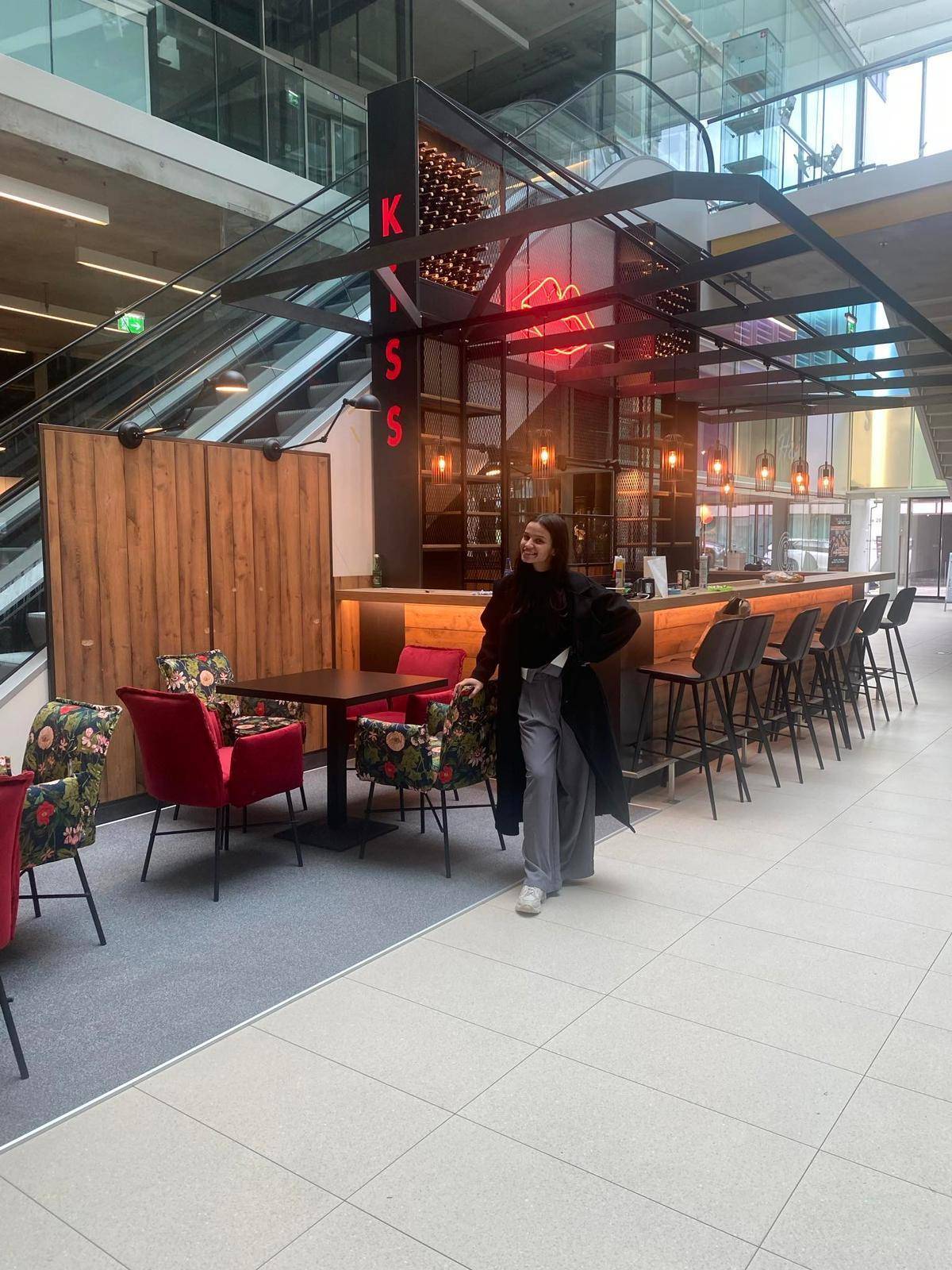 Moderne Bar-Lounge in einem großen Atrium mit Sitzgelegenheiten, Barhockern und Rolltreppen im Hintergrund.