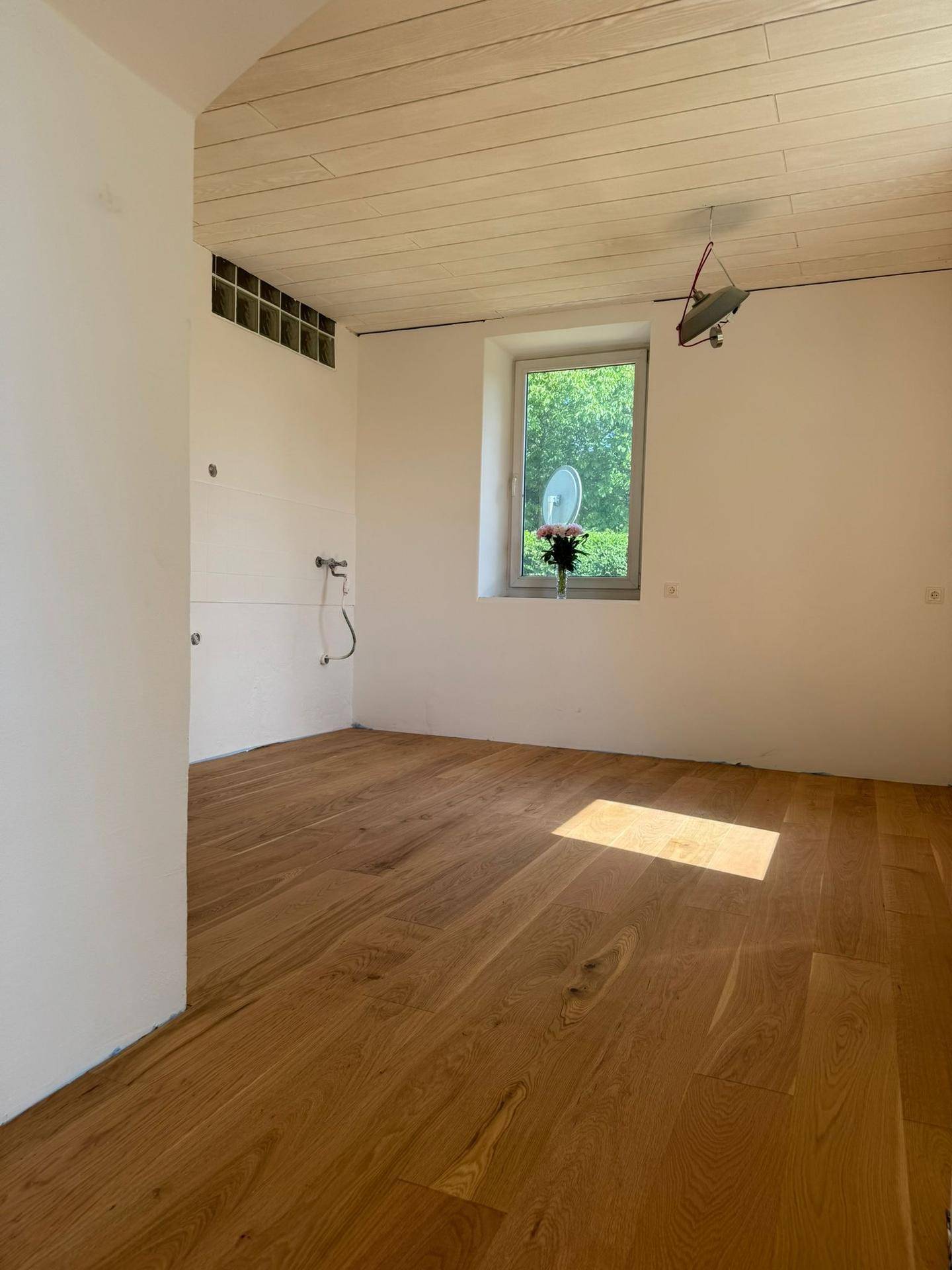 Heller Raum mit Holzparkettboden, Fenster und sichtbaren Anschlüssen an der Wand.