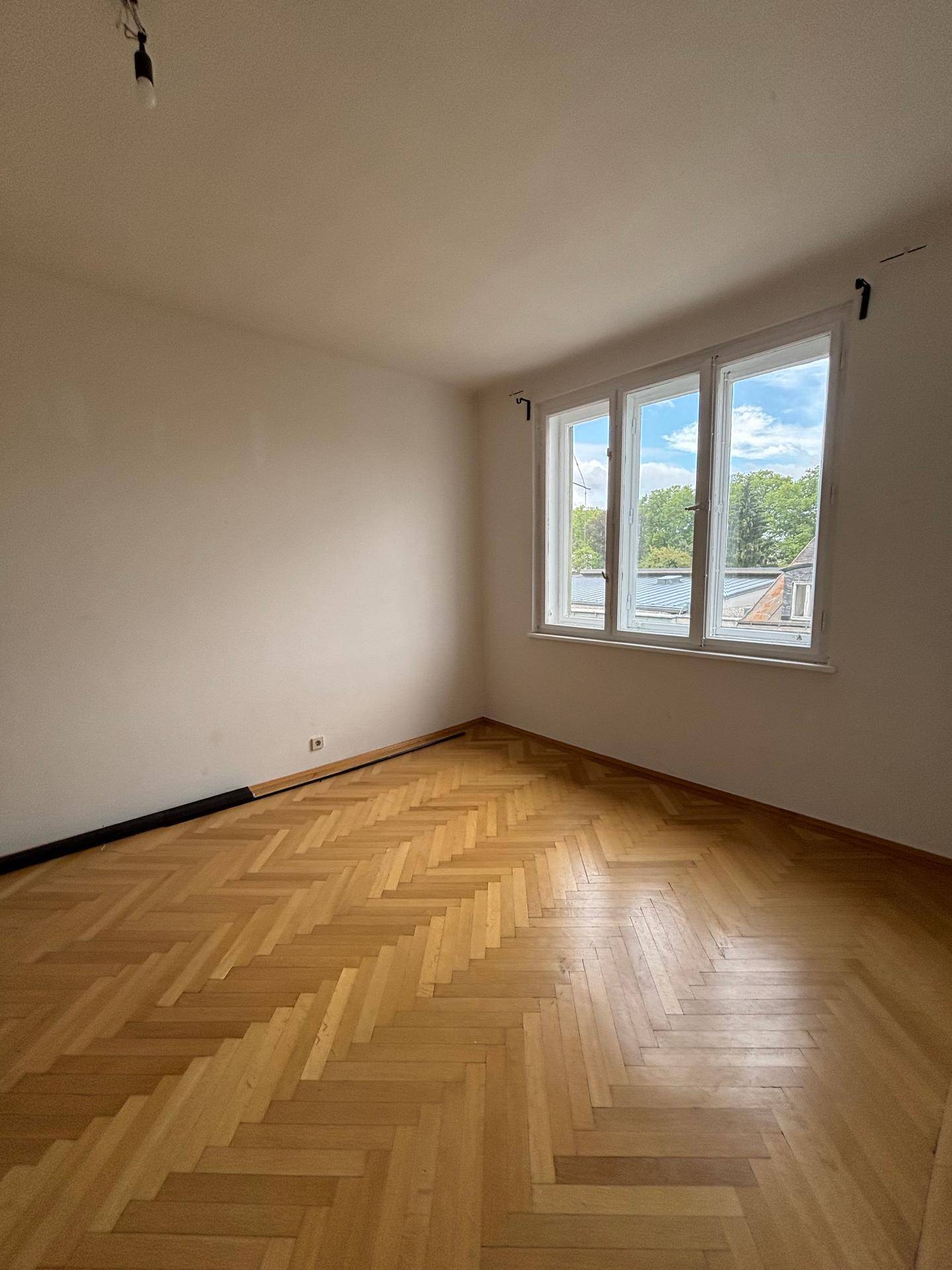 Helles Zimmer mit Fischgrätparkett und einem Fenster mit Blick ins Grüne und auf Dächer.