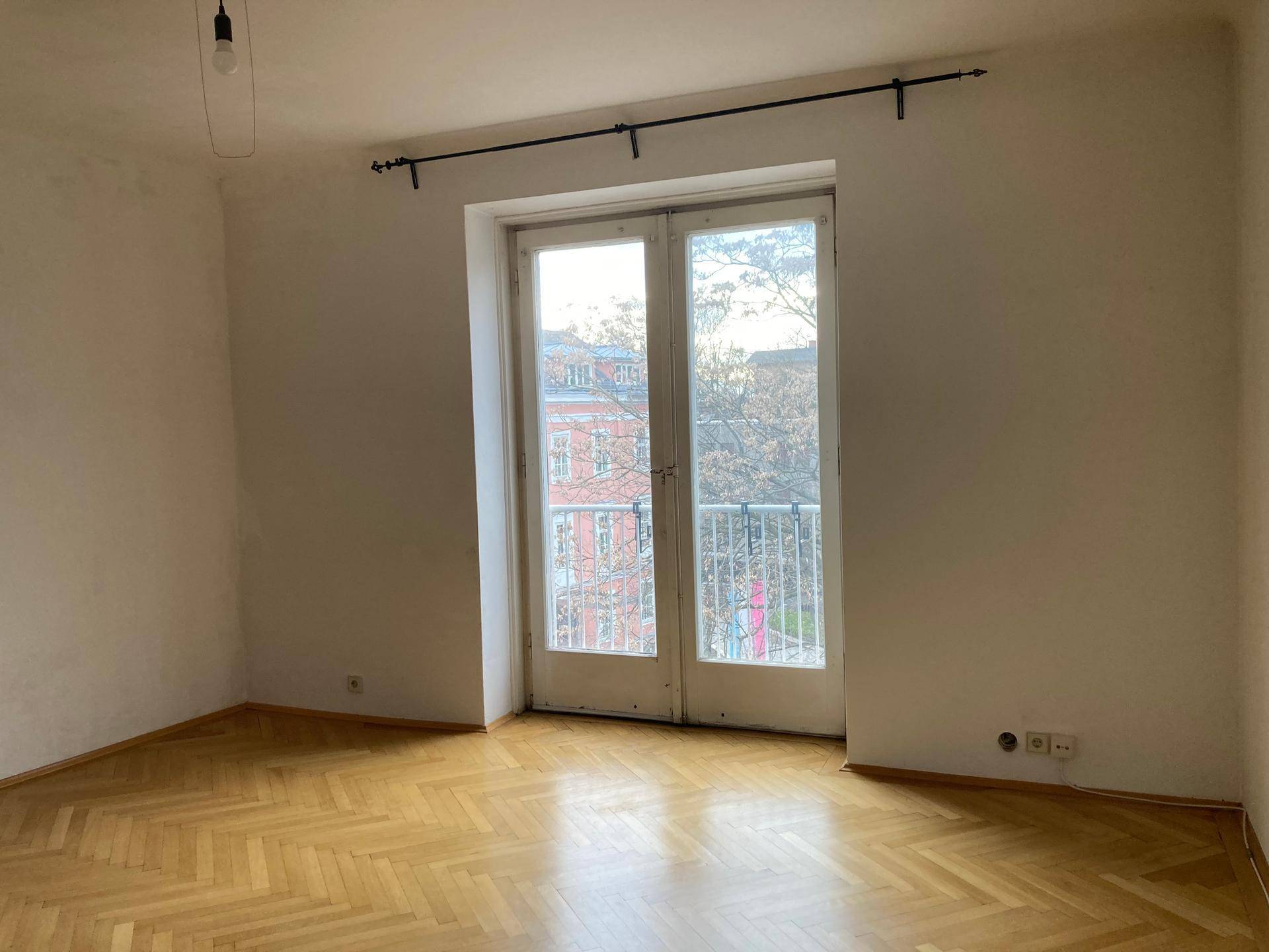Helles Zimmer mit Fischgrätparkett und einem französischen Balkon, der viel Tageslicht hereinlässt.