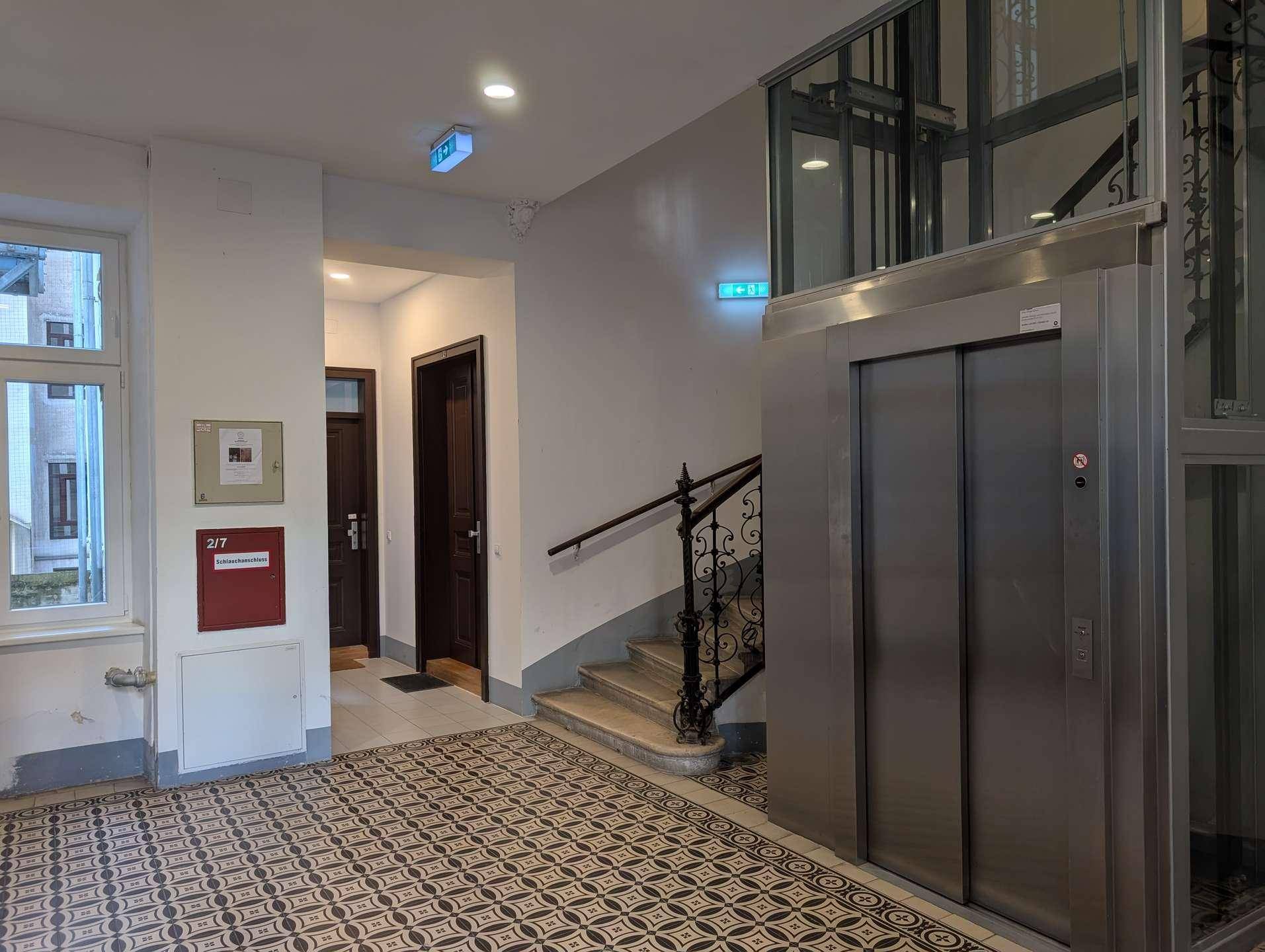 Geräumiger Flur im Gebäude mit Aufzug, Treppe und dekorativen Bodenfliesen.