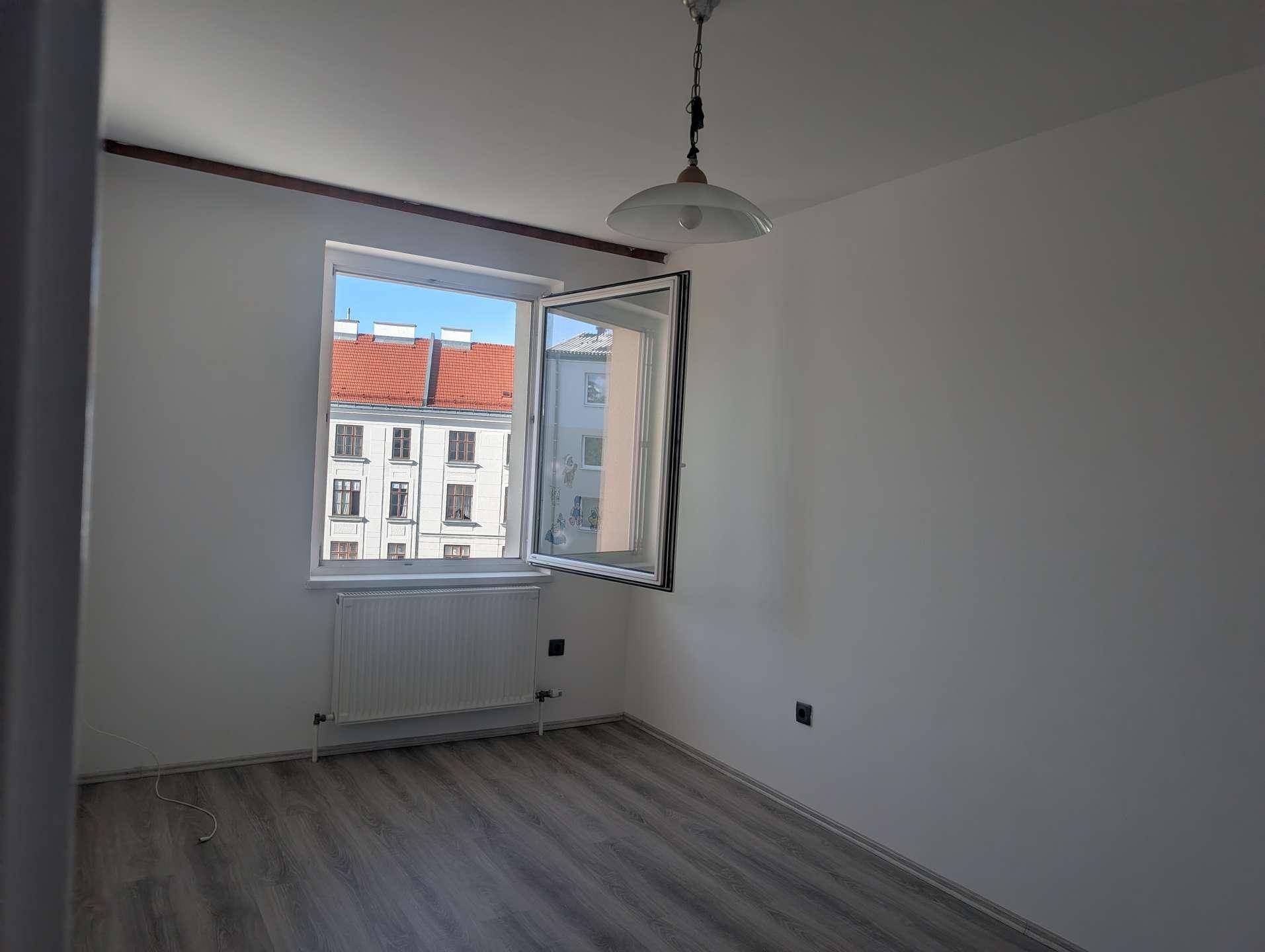 Leeres Zimmer mit grauem Laminatboden und geöffnetem Fenster, das Tageslicht hereinlässt.
