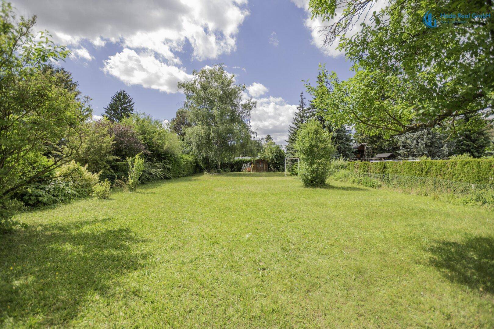 Ausgedehnter Gartenbereich mit viel Grün, Bäumen und offener Rasenfläche unter blauem Himmel.