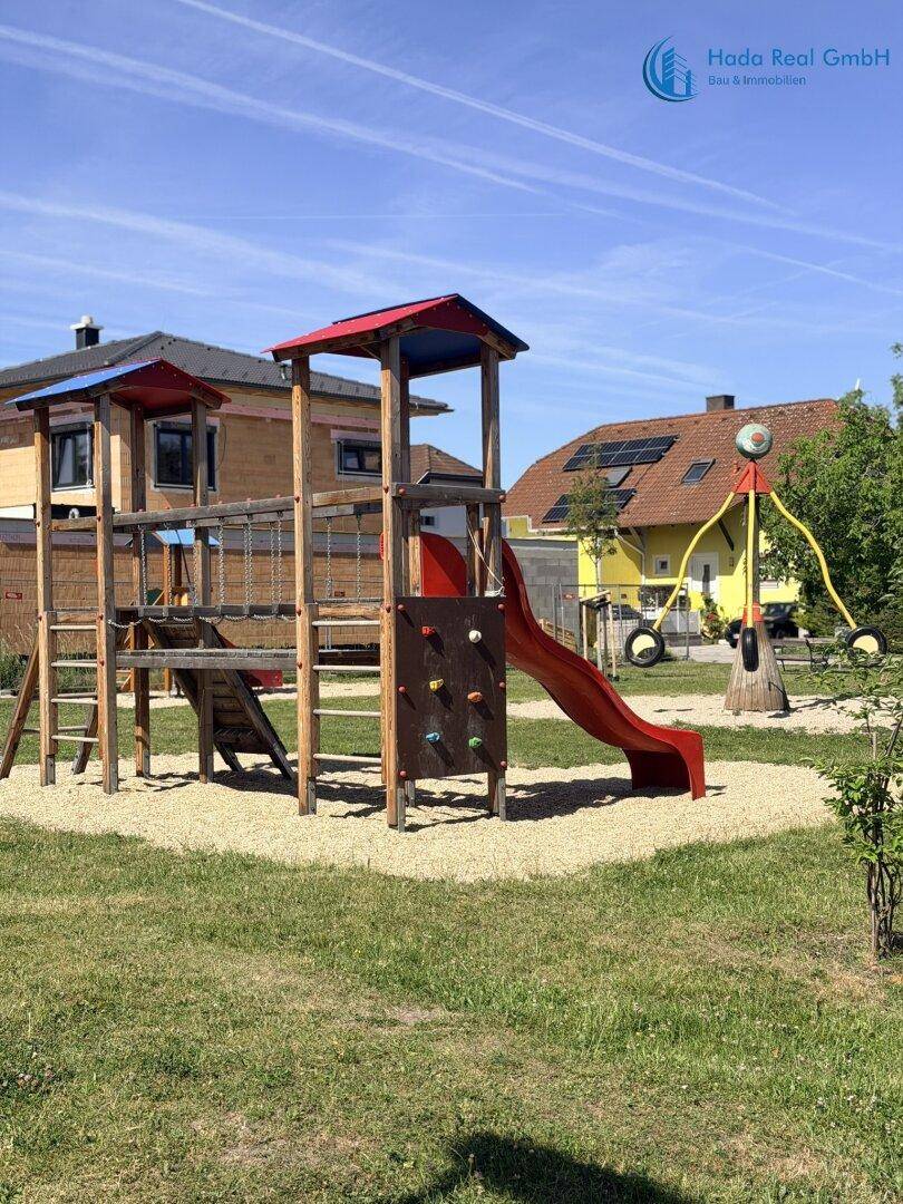 Öffentlicher Spielplatz mit Holzspielgeräten, Rutsche und Sandfläche, umgeben von Wohnhäusern.