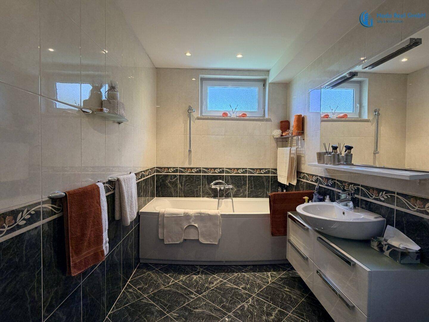 Badezimmer mit Badewanne, modernem Waschbecken und dunklen Fliesen, beleuchtet durch zwei kleine Fenster.