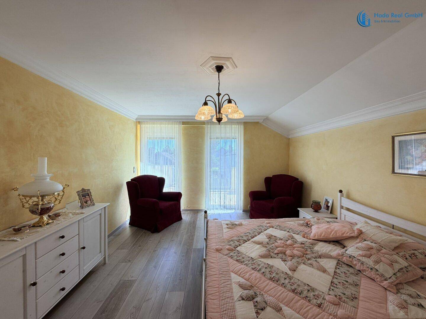 Helles Schlafzimmer mit Bett, Kommode und zwei Sesseln am Fenster, ausgestattet mit Holzboden.