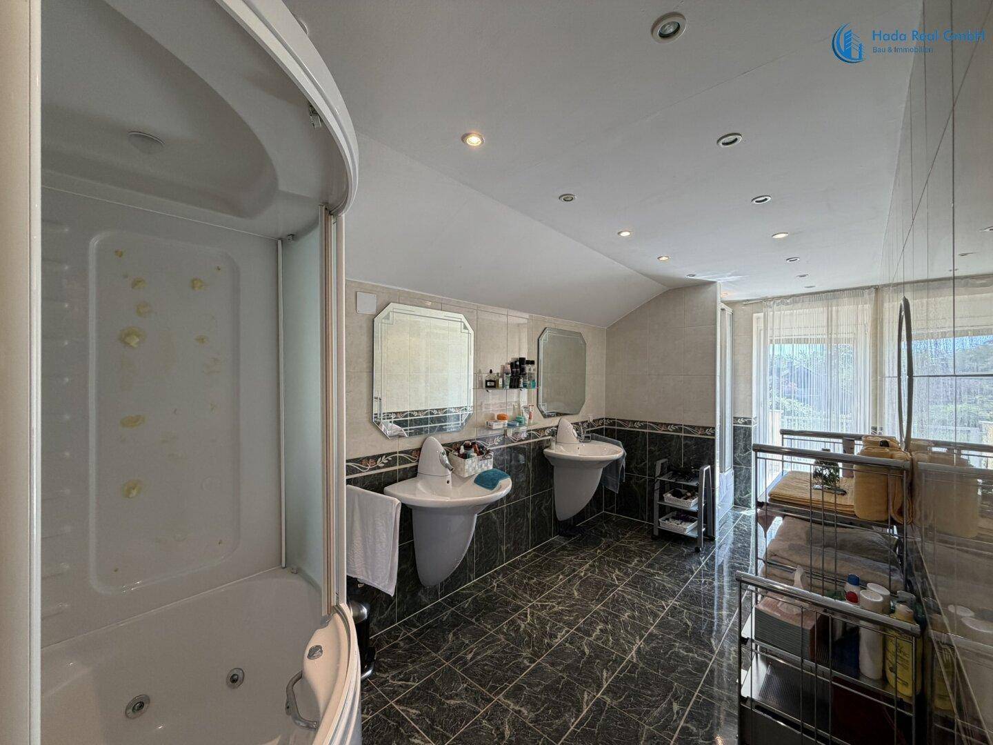 Geräumiges Badezimmer mit Eck-Whirlpool, Doppelwaschbecken und dunklen Fliesen unter einer Dachschräge.