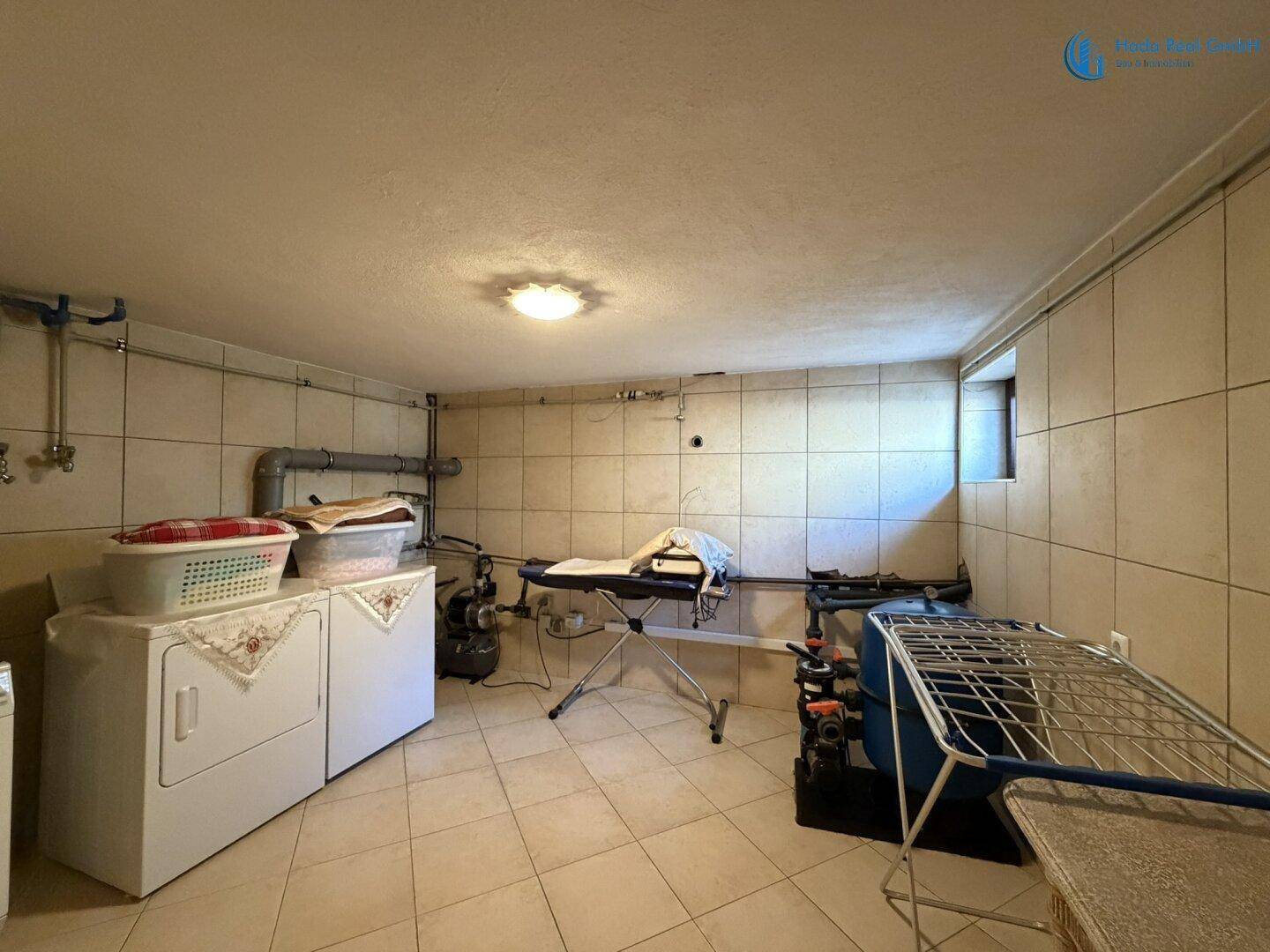 Gefliester Hauswirtschaftsraum im Keller mit Waschmaschine, Trockner und Wäscheständer.