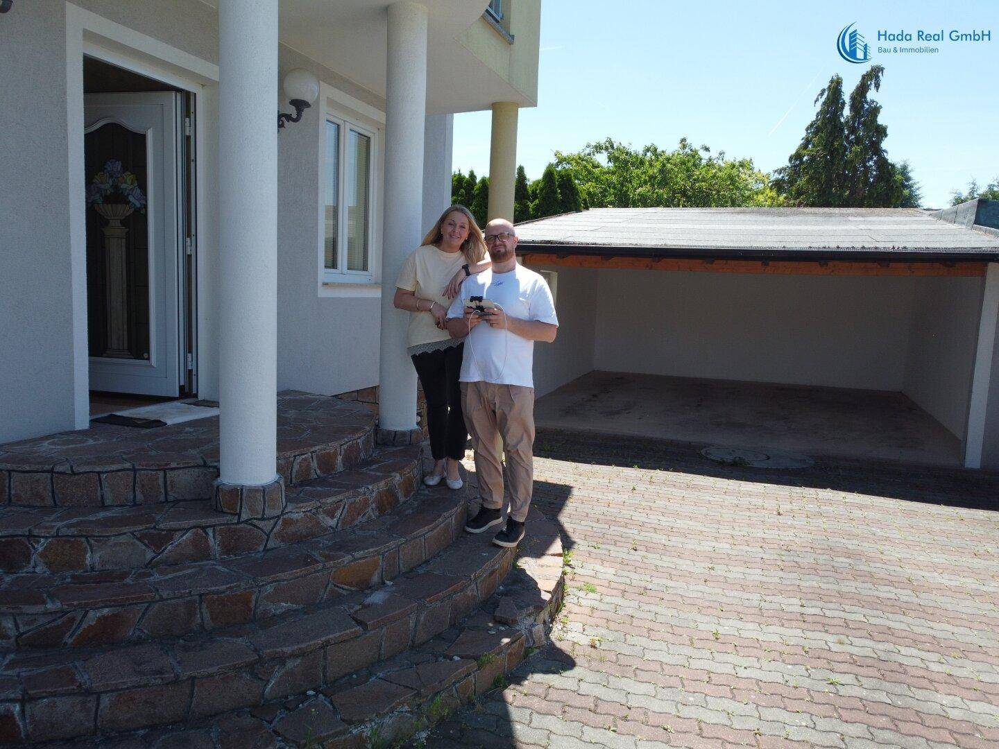 Zwei Personen stehen vor dem Eingang eines Hauses mit Steintreppe und einem Carport im Hintergrund.