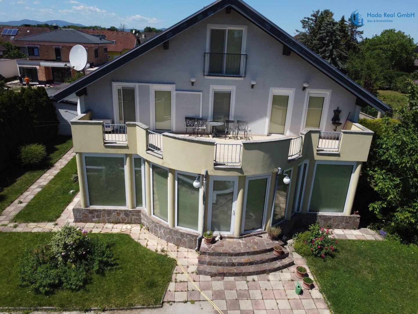Eine erhöhte Ansicht der Hausrückseite mit Balkonen, Wintergarten und einem gepflegten Rasenbereich.