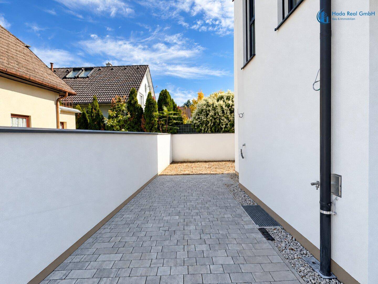 Gepflasterte Zufahrt neben dem Haus mit weißer Mauer und Blick auf Nachbargebäude.
