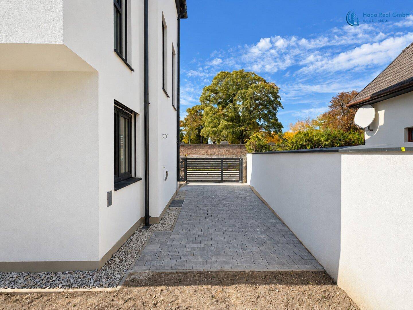 Gepflasterte Zufahrt zum Grundstück mit Tor und Blick auf Bäume im Hintergrund.