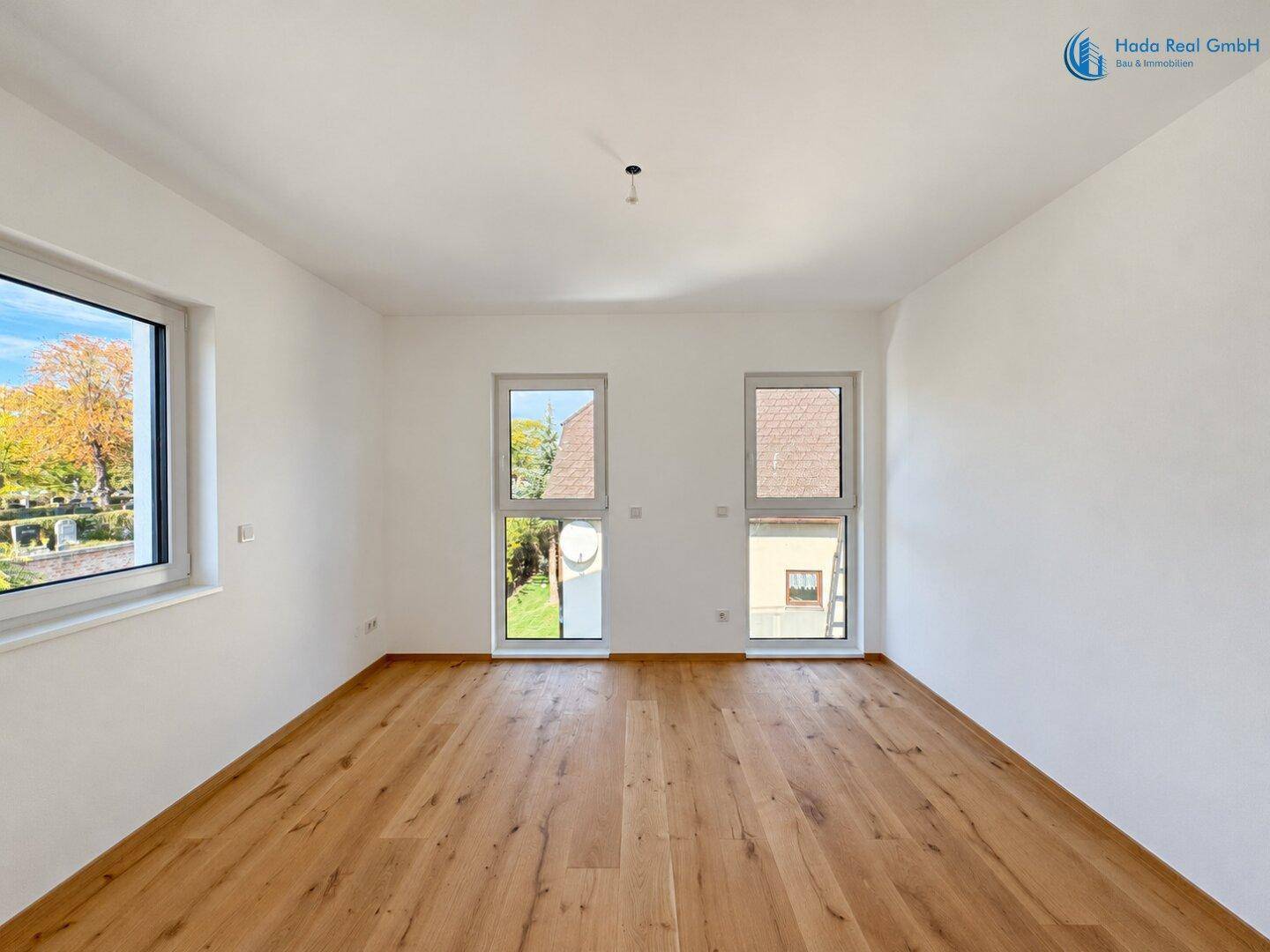 Freundliches Zimmer mit Holzboden und zwei hohen Fenstern, die einen Blick ins Grüne bieten.
