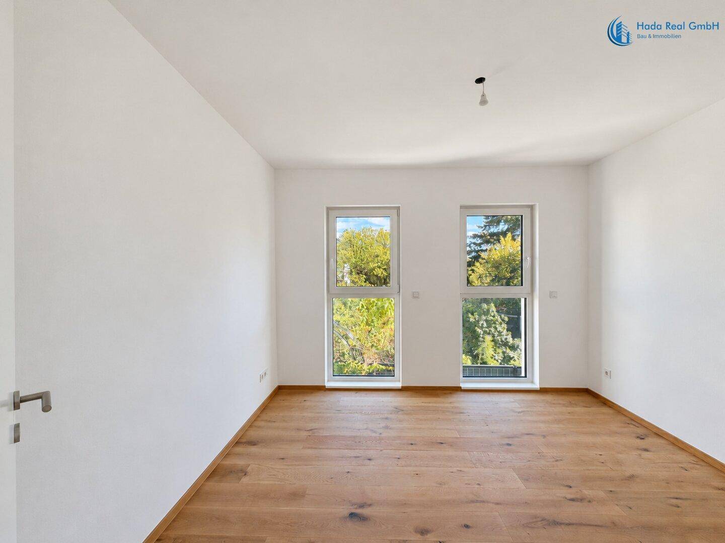 Helles Zimmer mit Holzboden und zwei Fenstern, die einen schönen Blick ins Grüne bieten.
