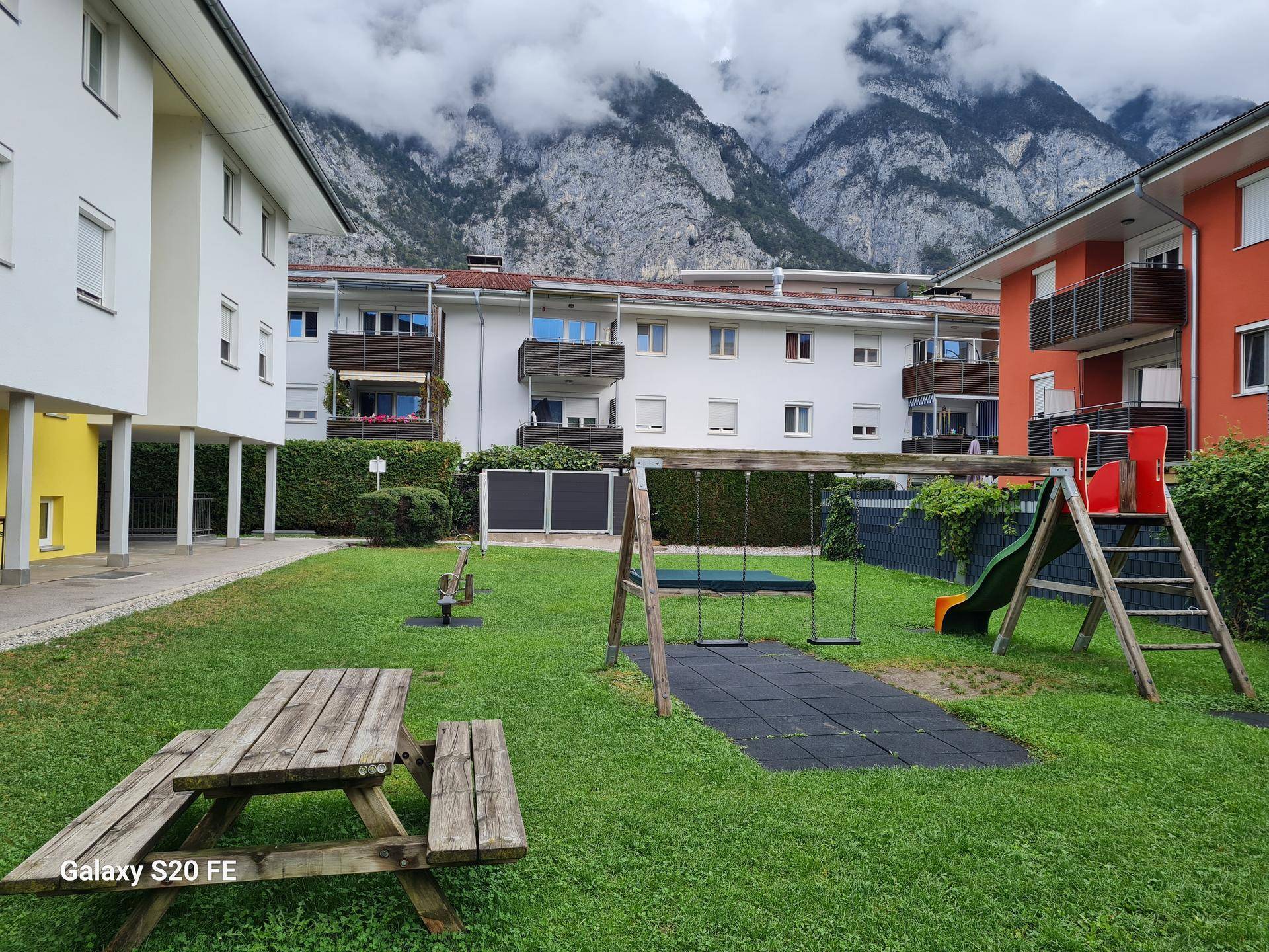 Gemeinschaftlicher Spielplatz mit Schaukeln und Rutsche, umgeben von Wohngebäuden und Bergen.
