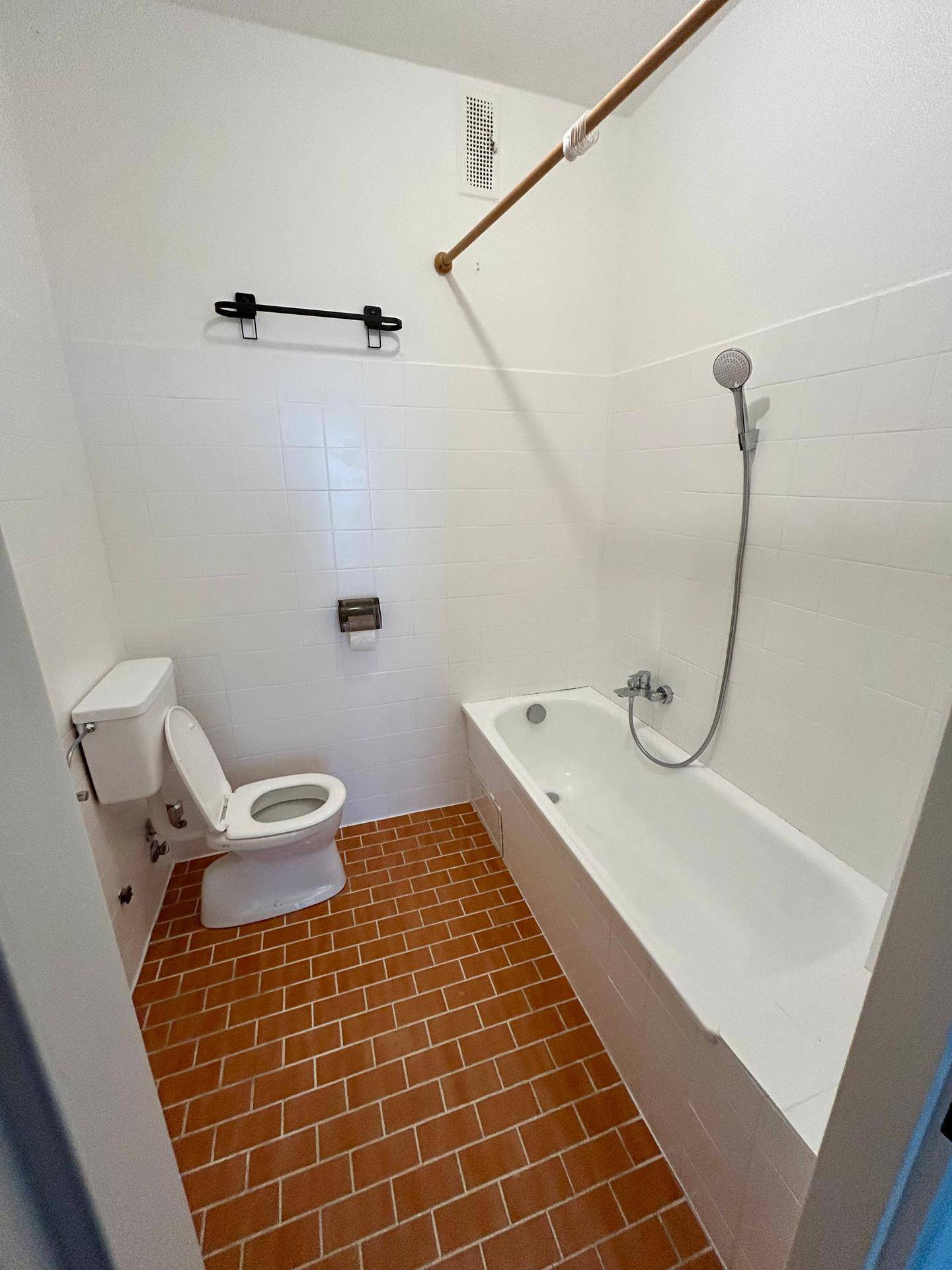 Badezimmer mit weißen Fliesen, Terrakotta-Bodenfliesen, Badewanne mit Duschfunktion und Toilette.