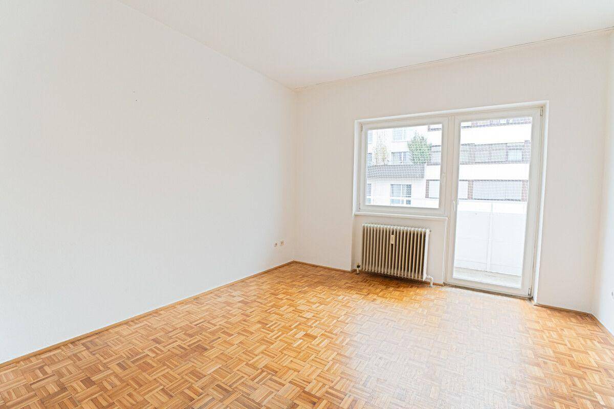 Geräumiges Zimmer mit Parkettboden, weiß gestrichenen Wänden und Zugang zu einem Balkon.