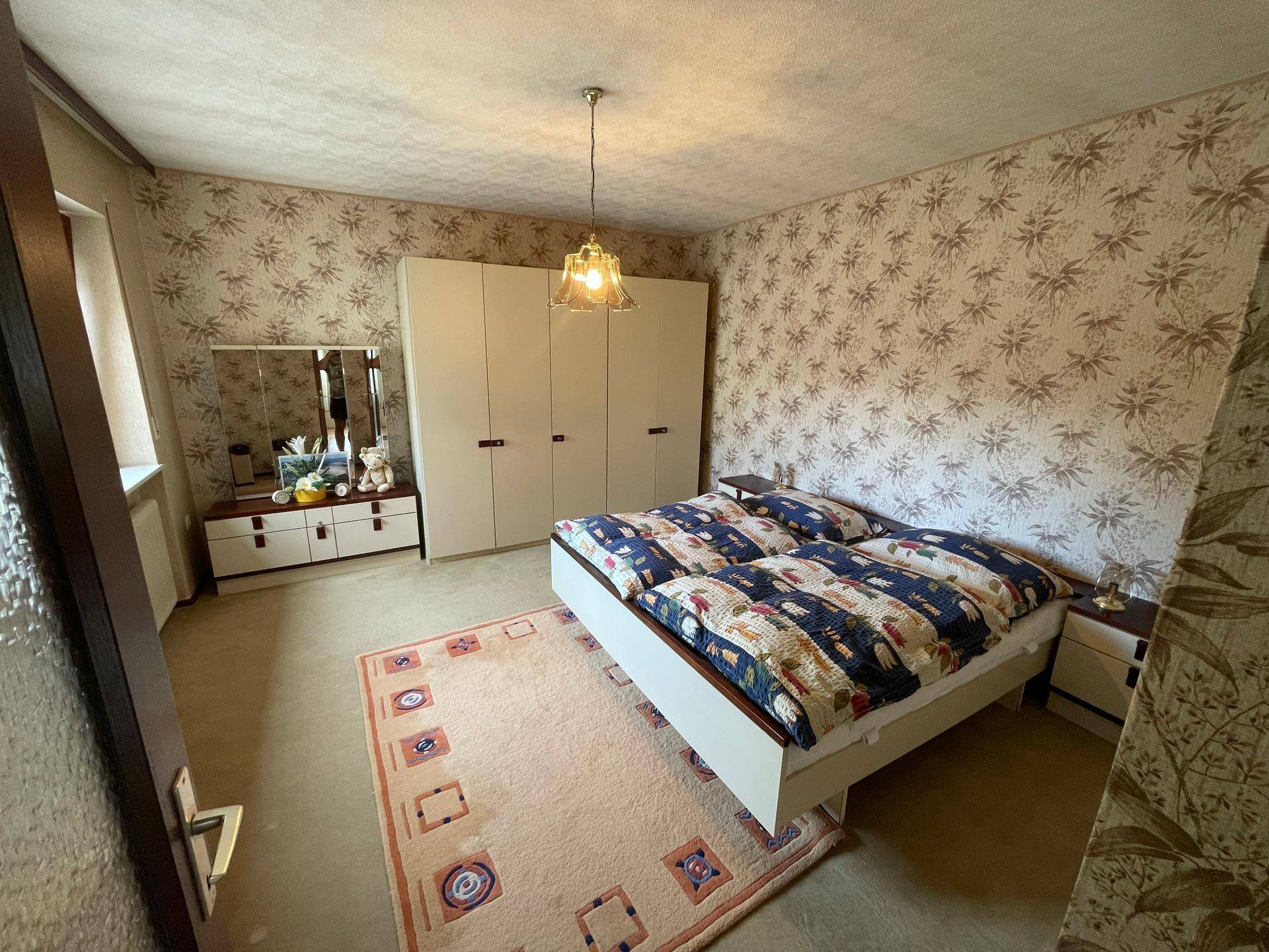 Schlafzimmer mit gemusterten Tapeten, Doppelbett und großem Kleiderschrank, bietet ausreichend Stauraum.