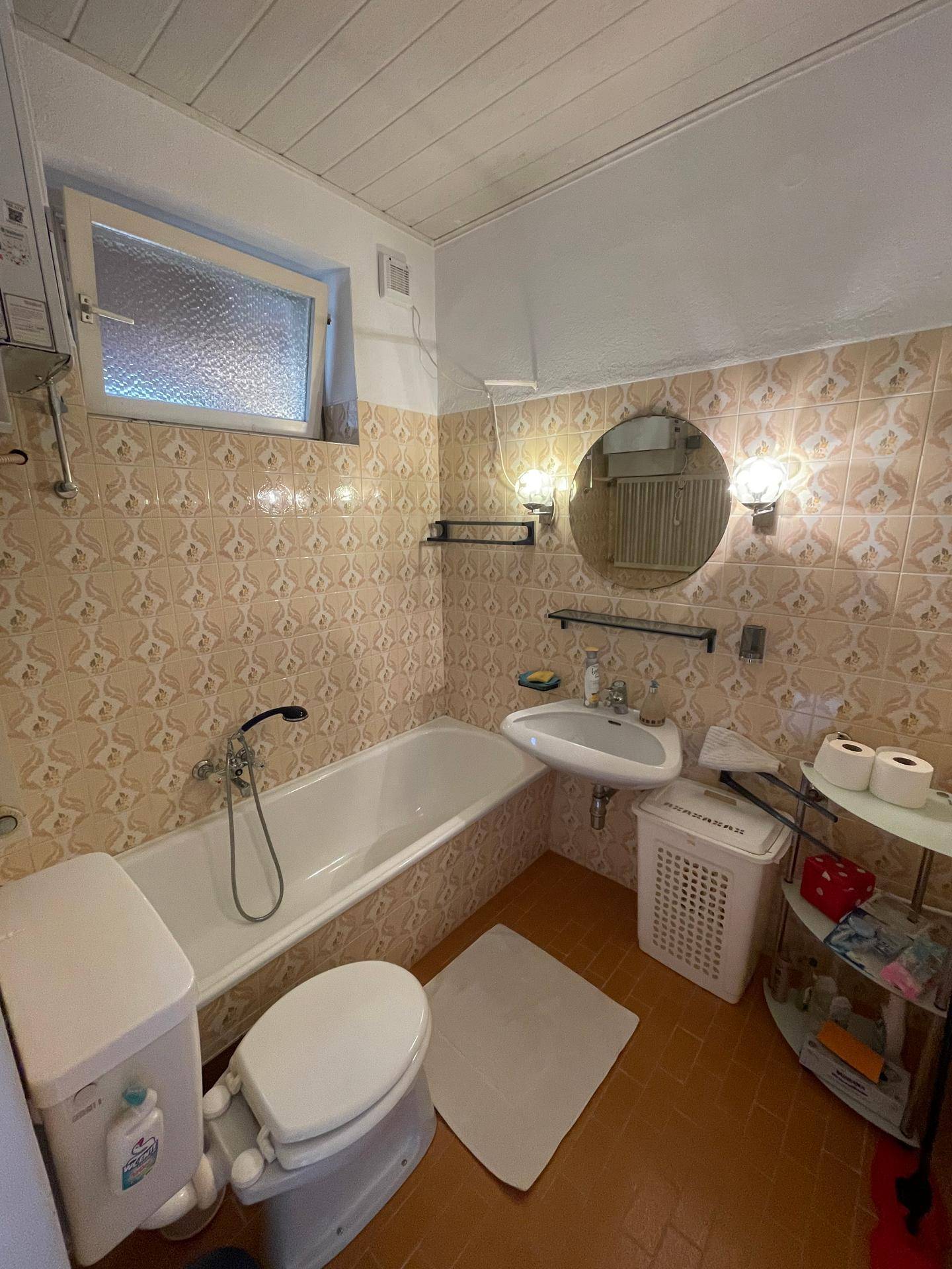 Badezimmer mit Badewanne, Waschbecken und Fenster, ausgestattet mit hellbraunen Fliesen und Holzdecke.